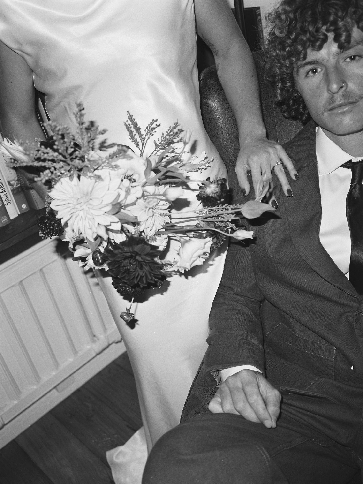 Black and white photo of bride in satin gown holding large floral bouquet beside groom in dark suit.