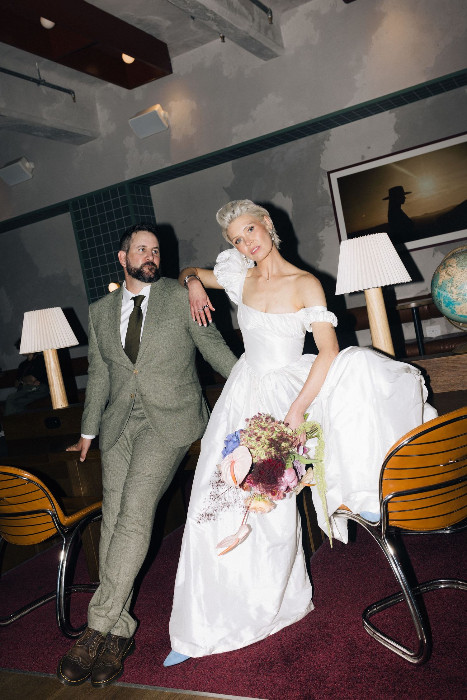 Bride in white off-shoulder gown holds bouquet beside groom in green suit at an indoor wedding venue.