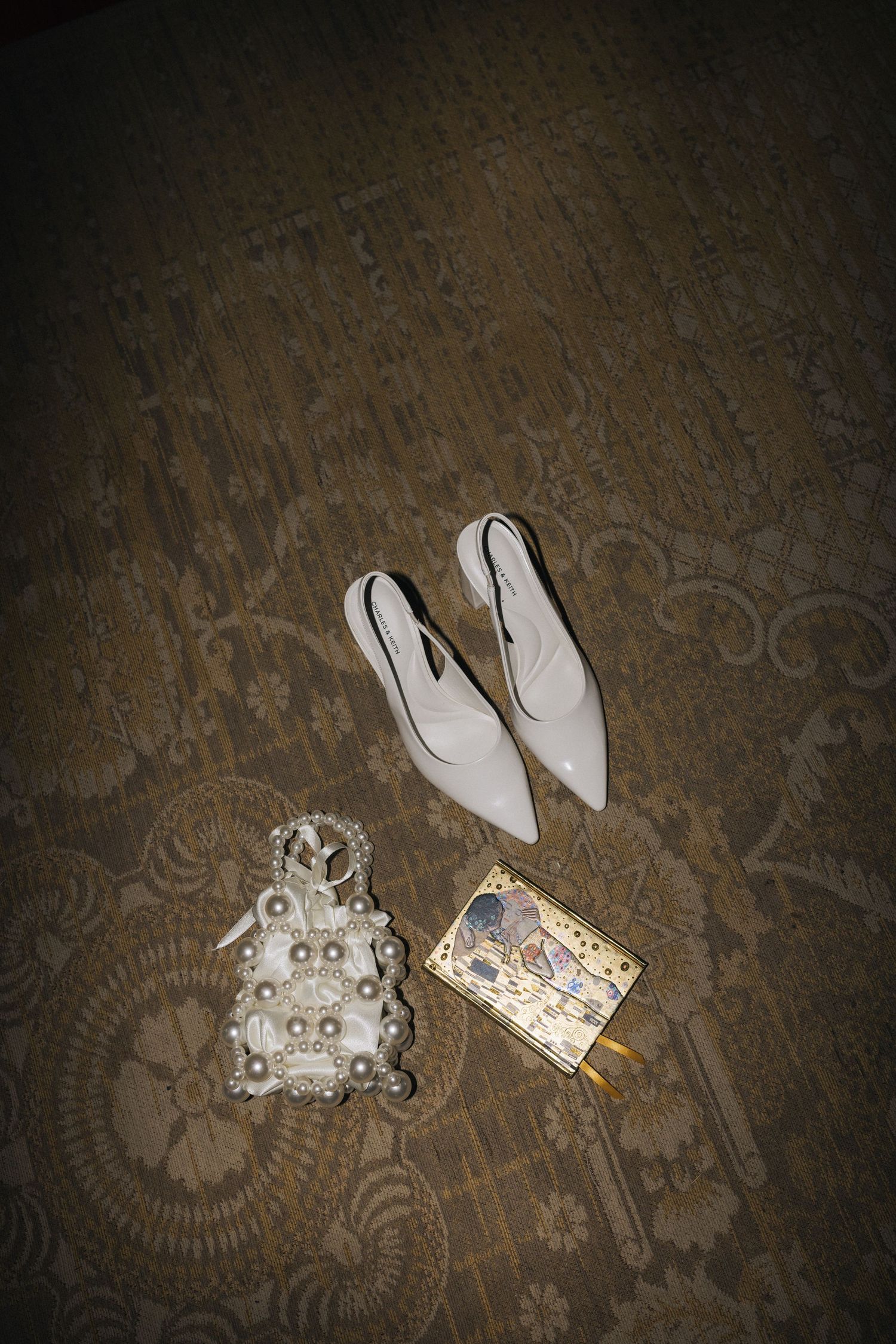 Flat lay of white bridal slingback heels, pearl bag, and decorative clutch on carpet.