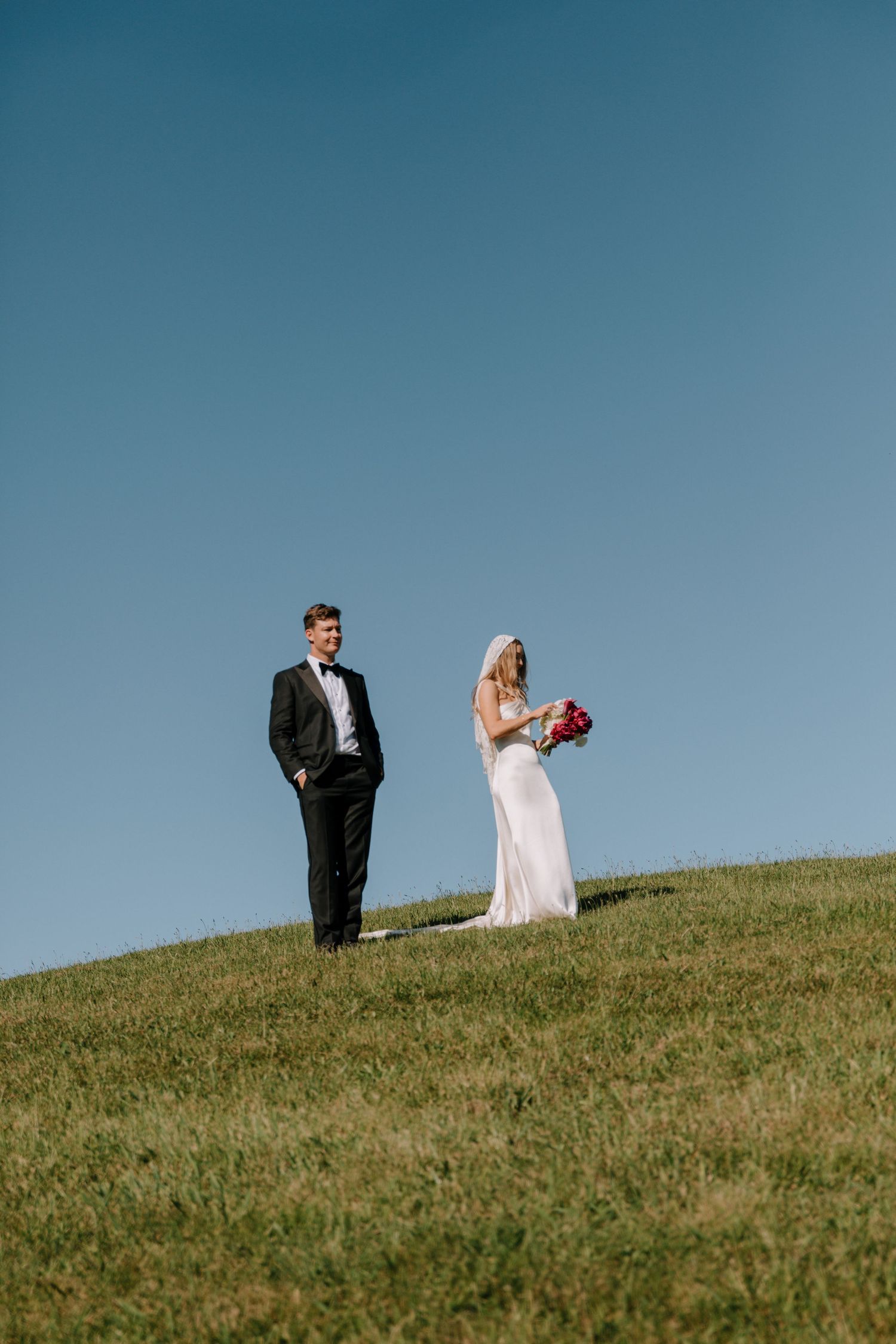 Bride and groom standing on green hillside against clear blue sky, bride holding red bouquet.