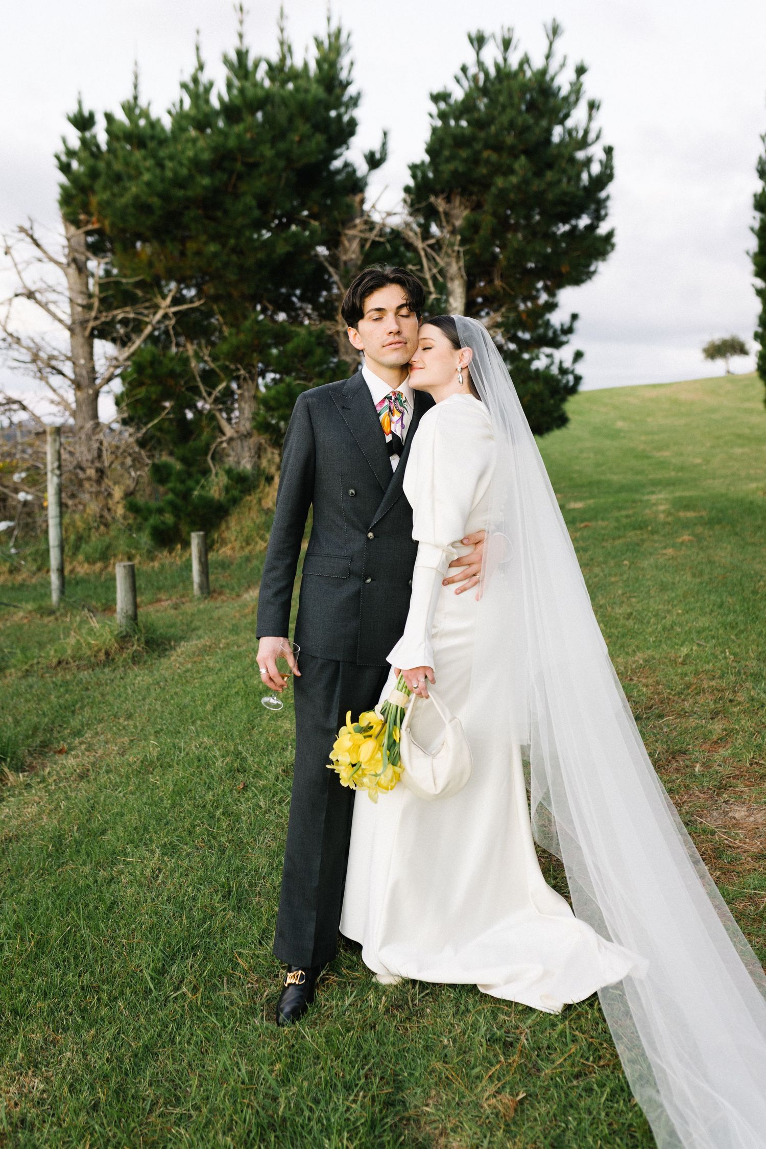 Bride in white long-sleeve gown with veil holds yellow bouquet beside groom in dark suit outdoors.