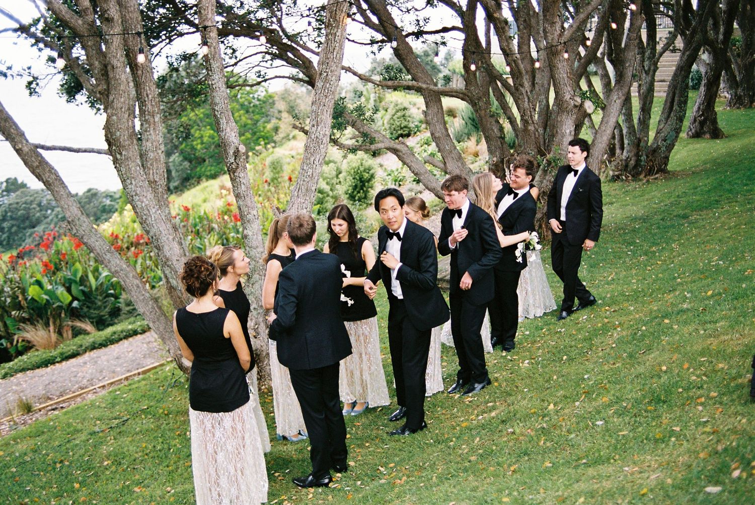Wedding party in formal attire socializes on a sloped green lawn among trees.