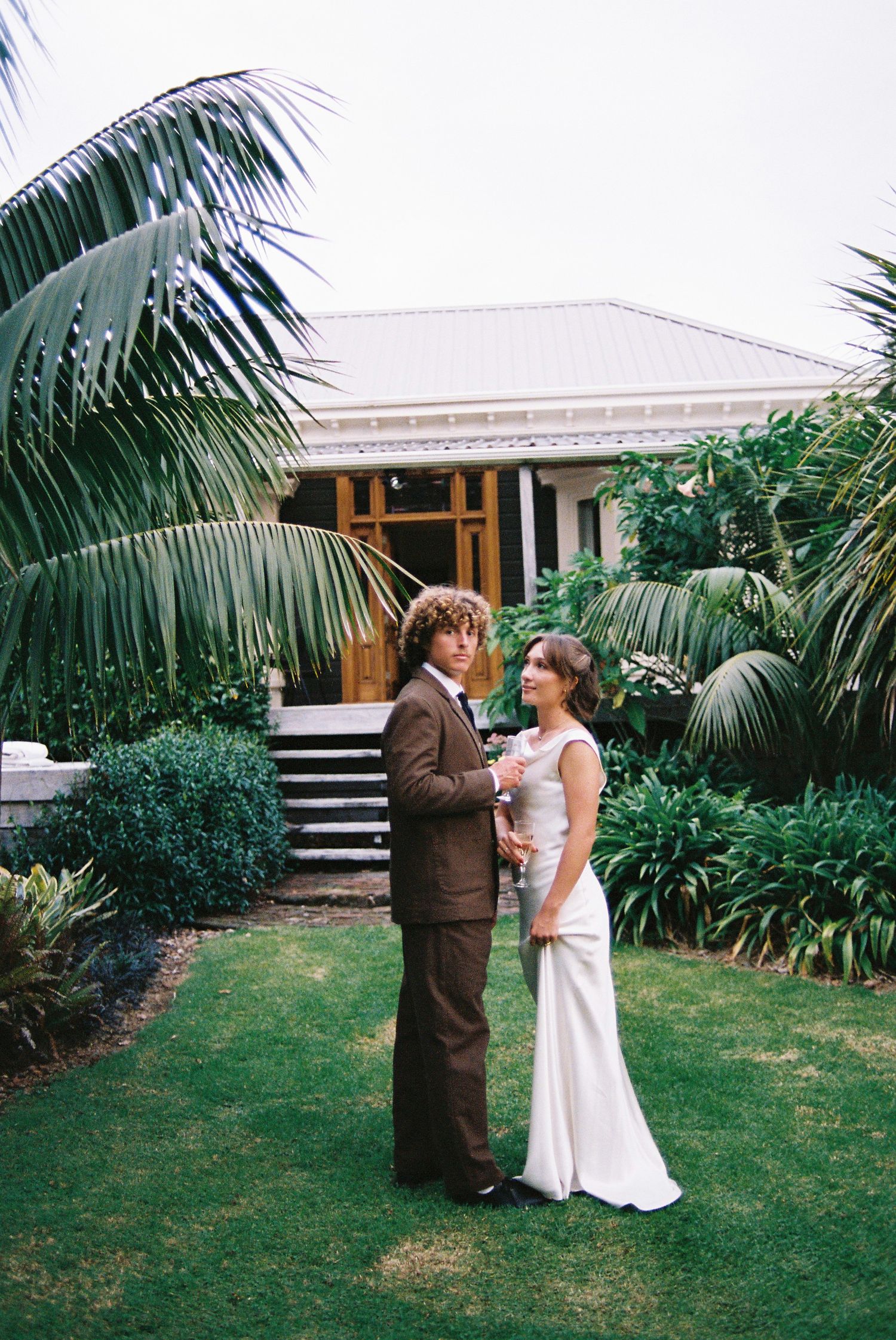 Bride and groom standing together on a lawn in front of a tropical garden and white colonial house.
