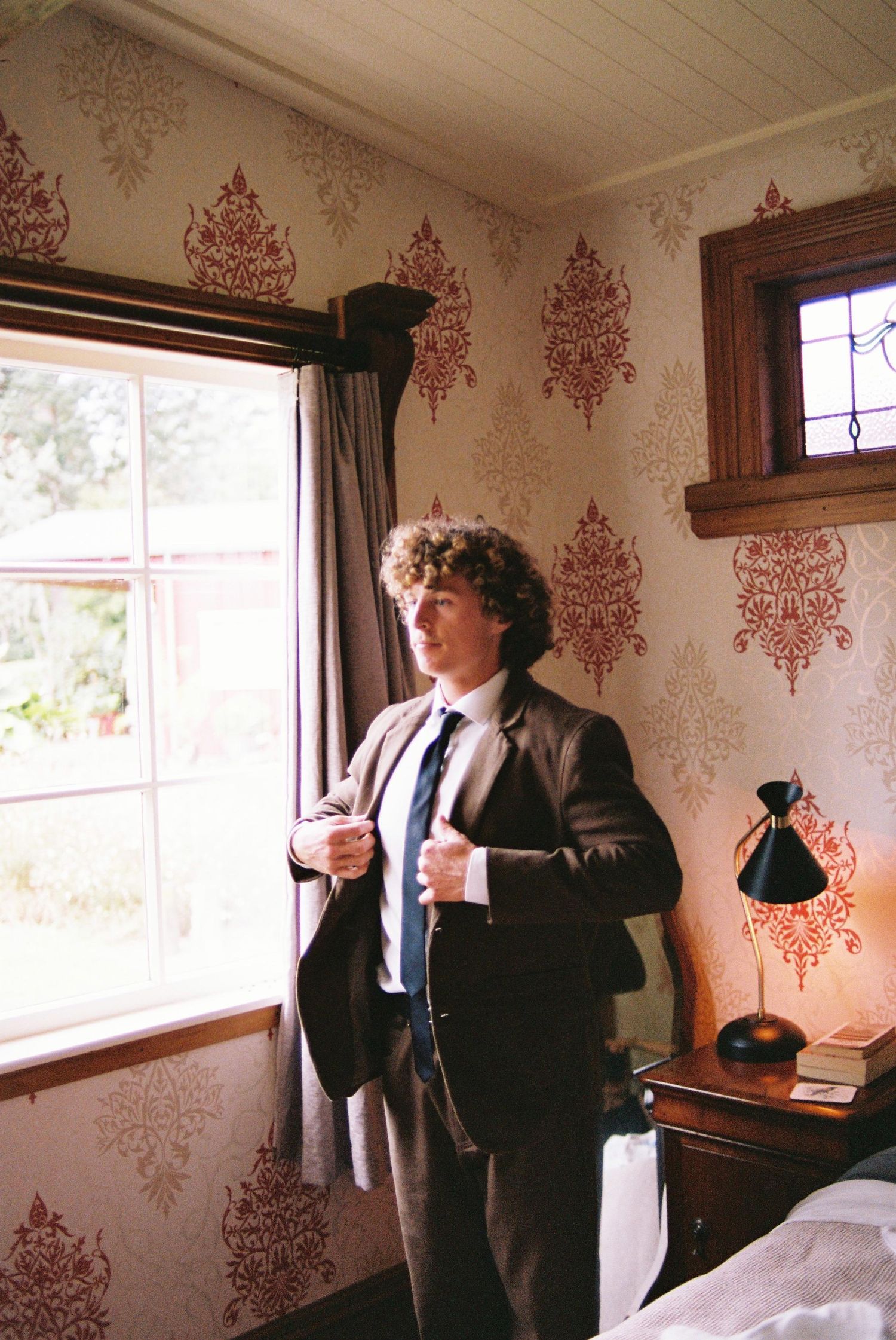 Curly-haired groom in brown suit adjusting jacket near window in room with ornate red damask wallpaper.