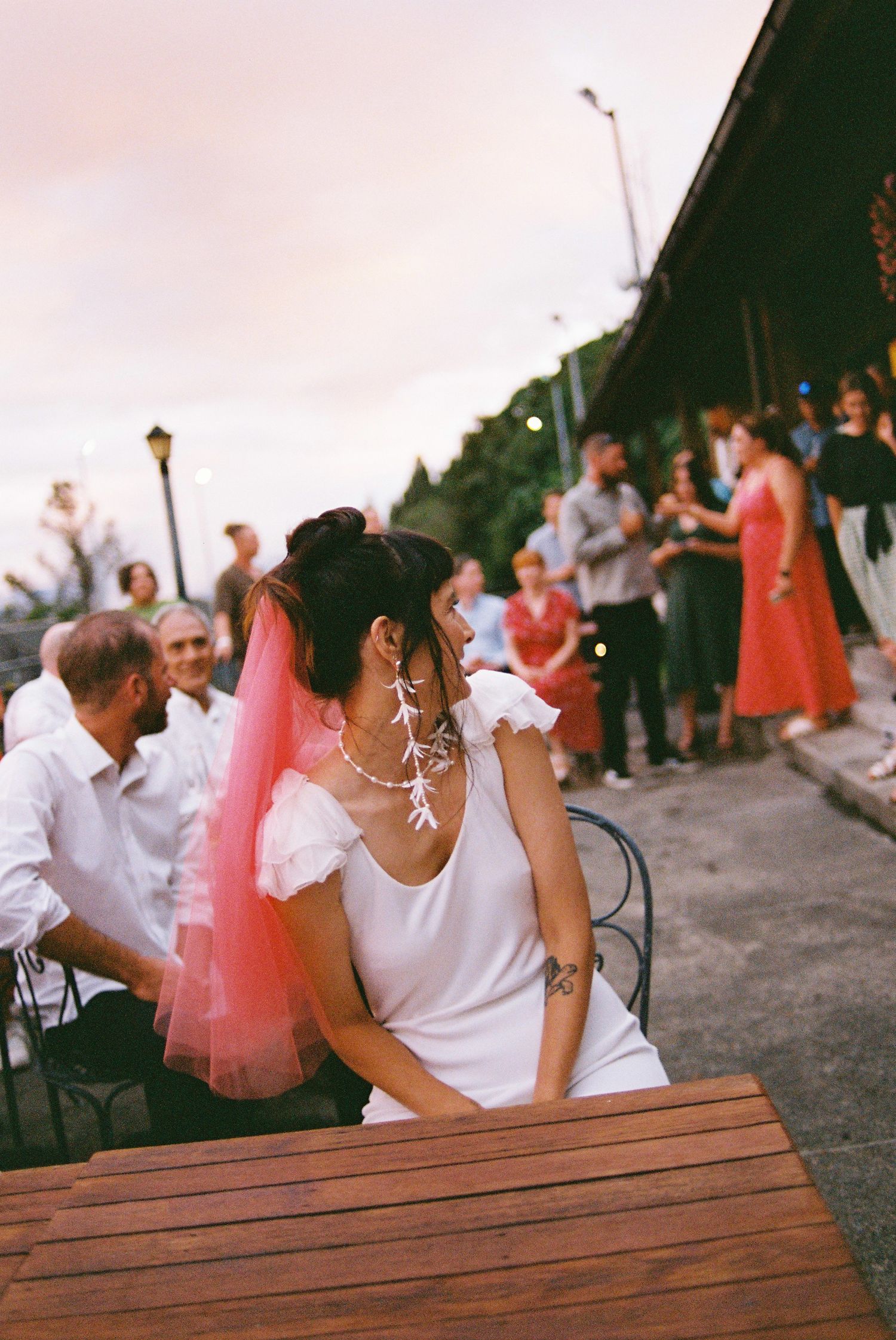 Bride in white dress with pink veil seated at outdoor table at dusk, guests mingling in background.