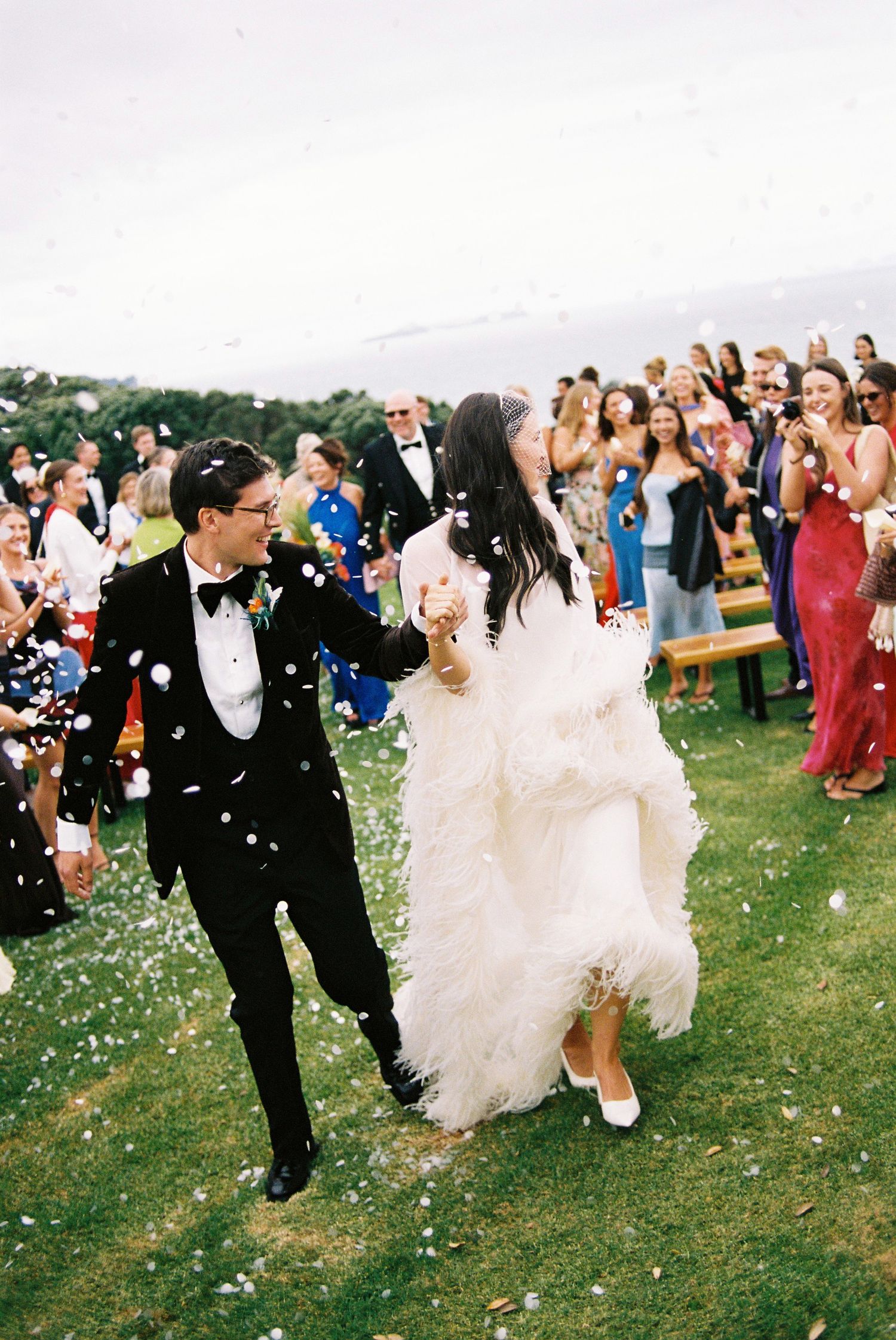 Bride and groom walk through confetti shower during outdoor wedding ceremony recessional.