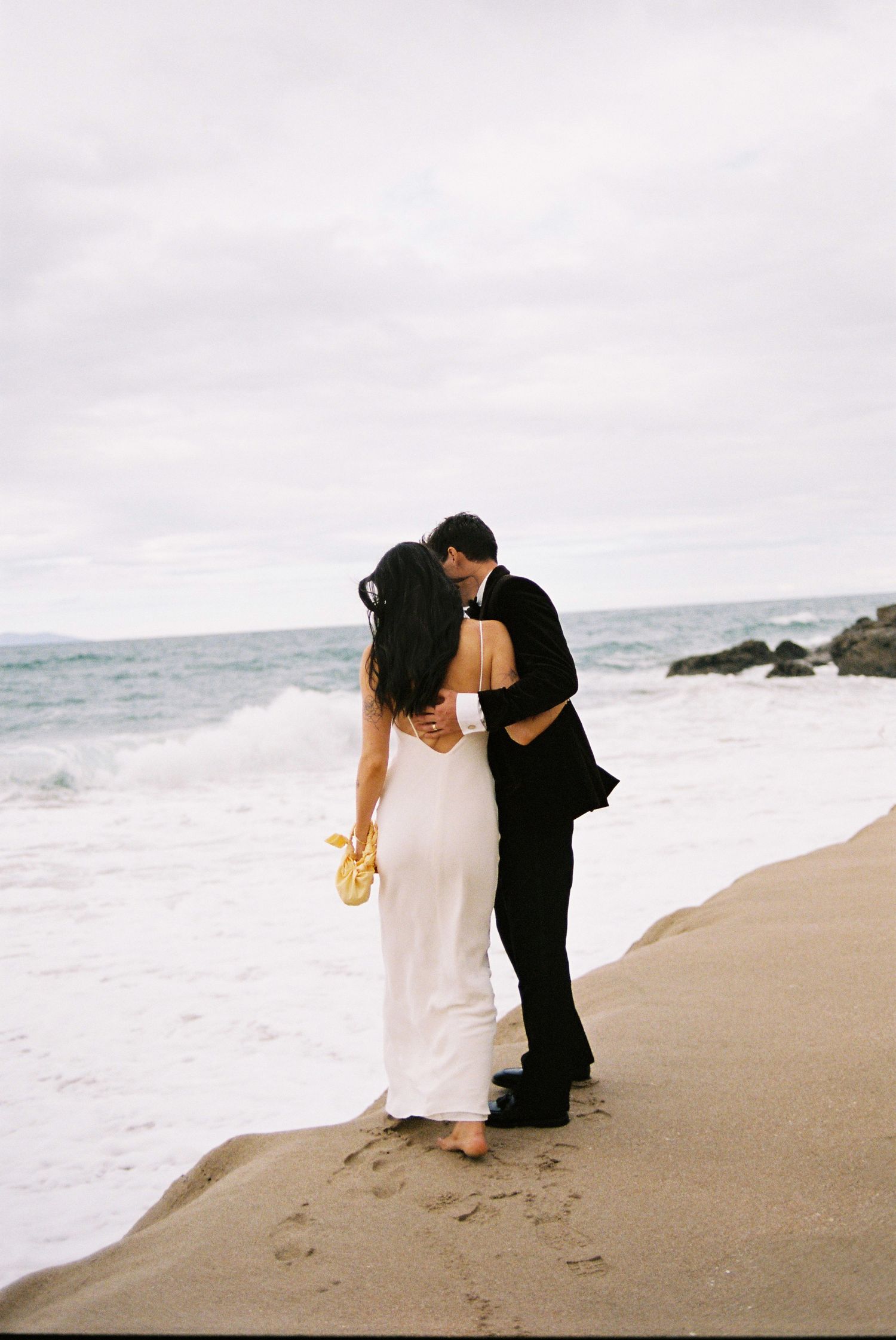 Bride in white slip gown and groom in black suit kissing on sandy beach with ocean waves behind them.