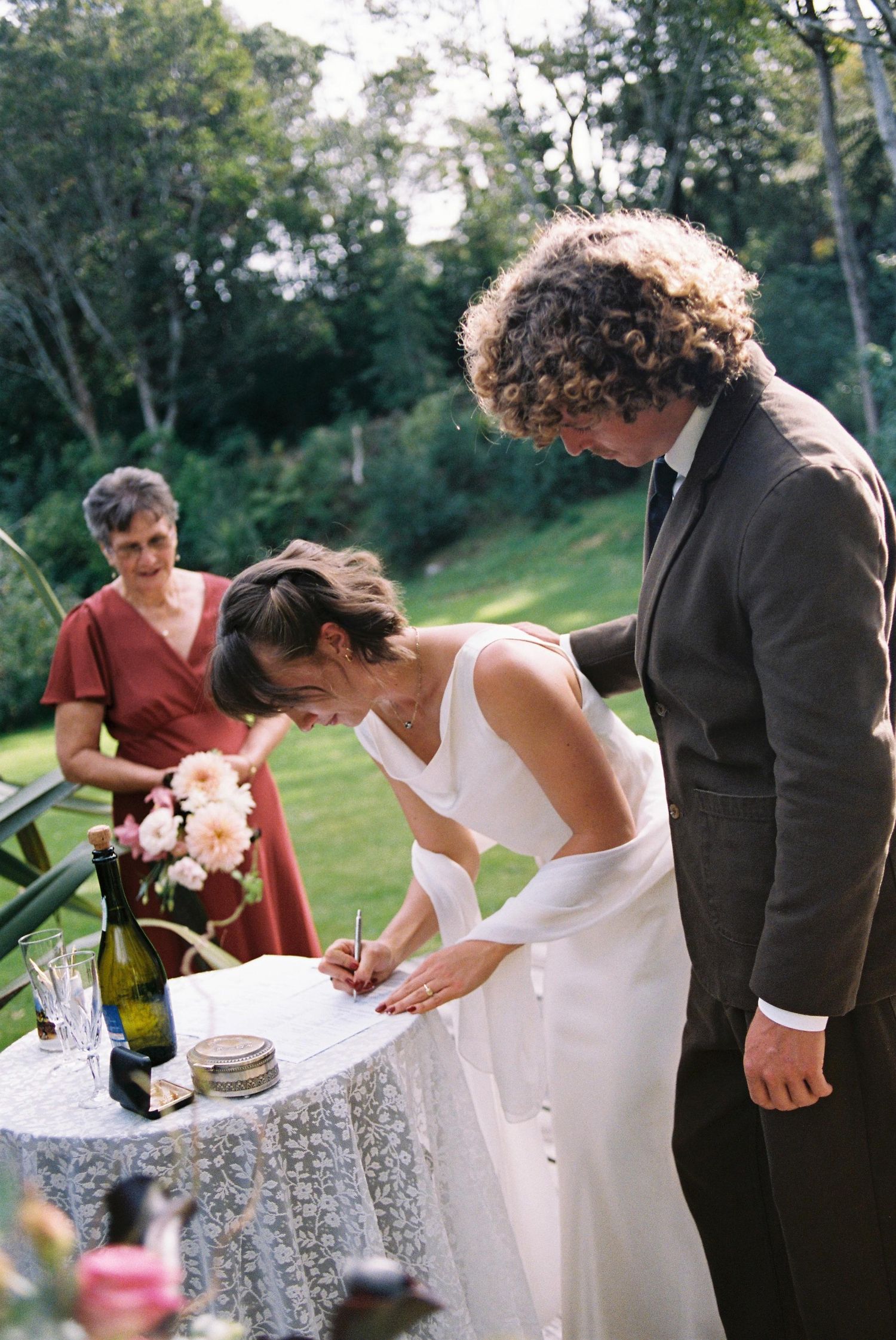 Bride signing wedding register at outdoor ceremony table with groom and officiant in green garden setting.