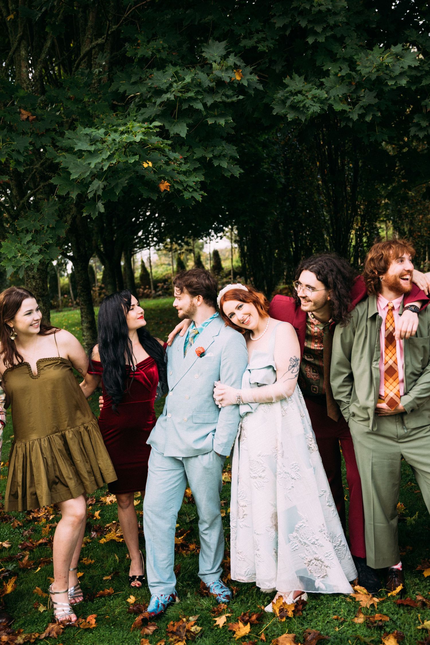 Wedding party in colorful vintage-inspired outfits laughing together among autumn trees.