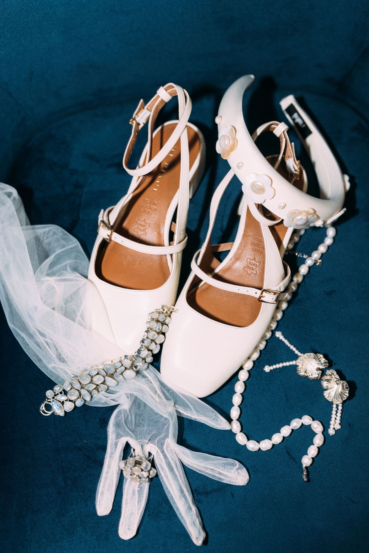 White bridal heels with floral details beside pearl jewelry and a veil on blue velvet.