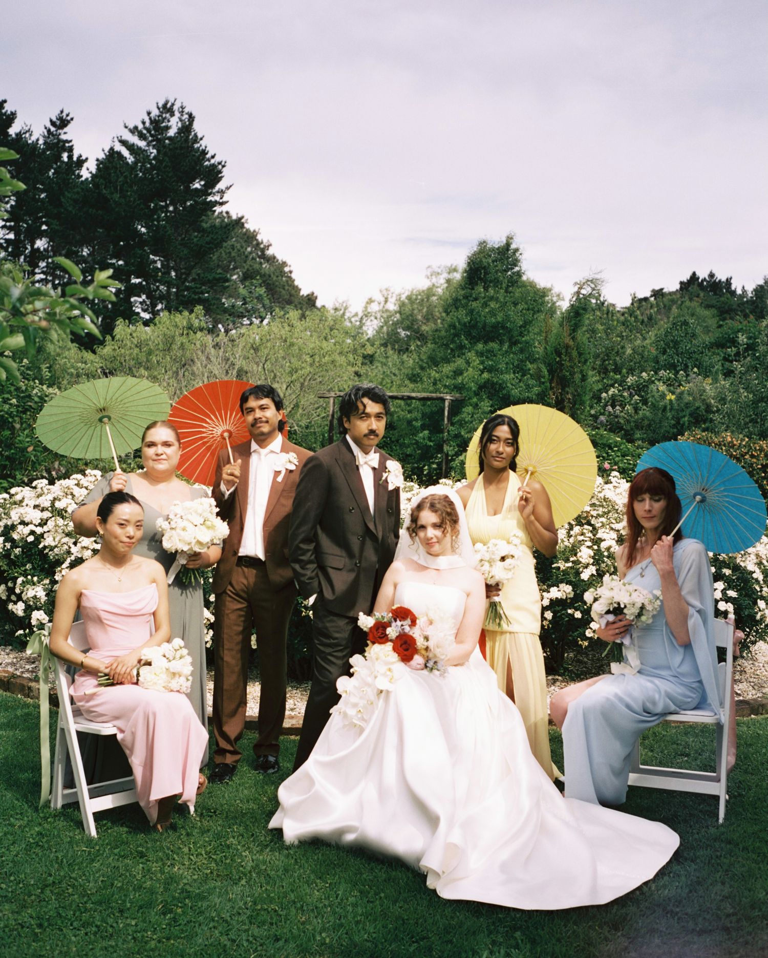 Wedding party poses with colorful parasols in garden with white flowers and greenery.