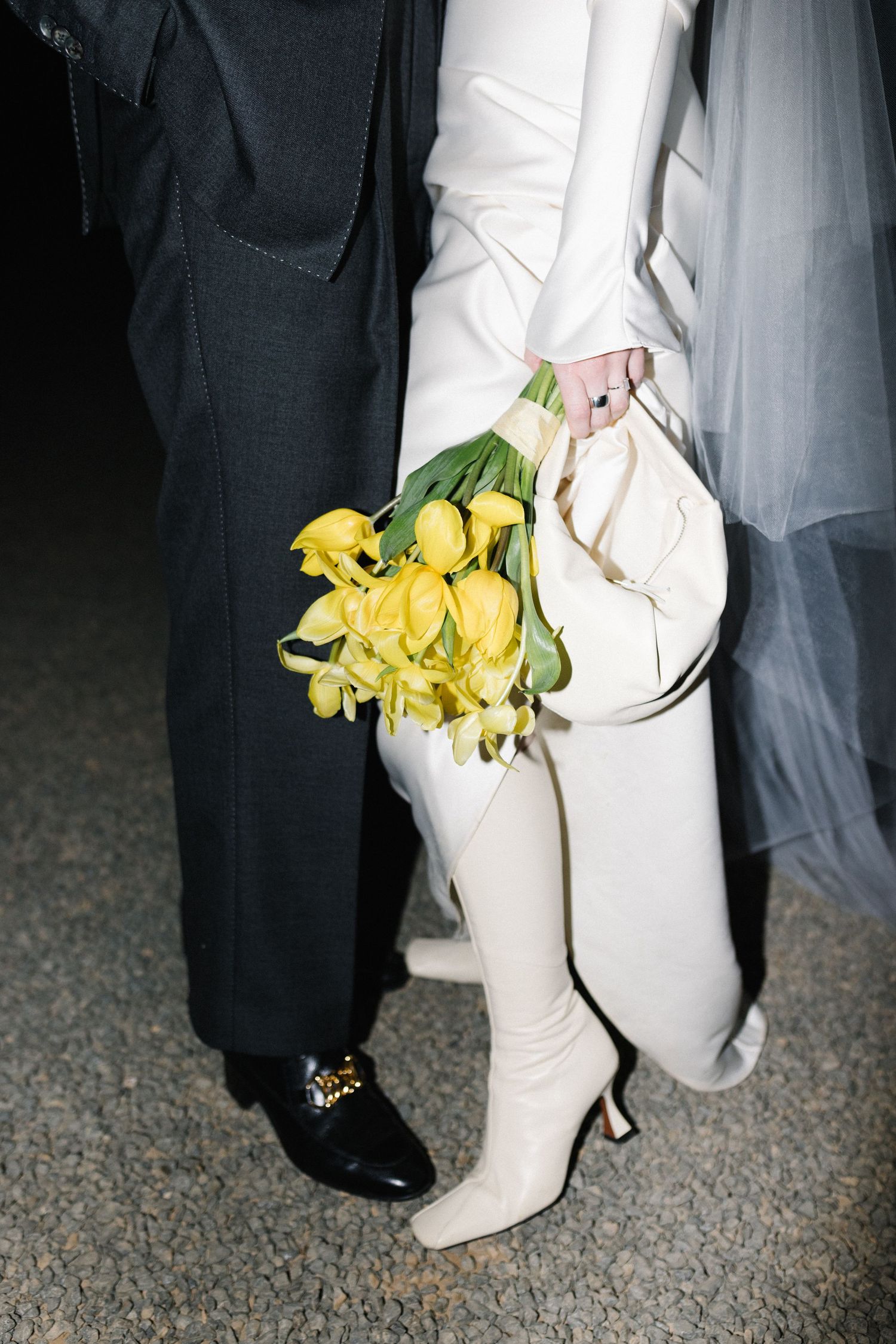 Close-up of bride in white boots holding yellow tulips beside groom in black suit and loafers.