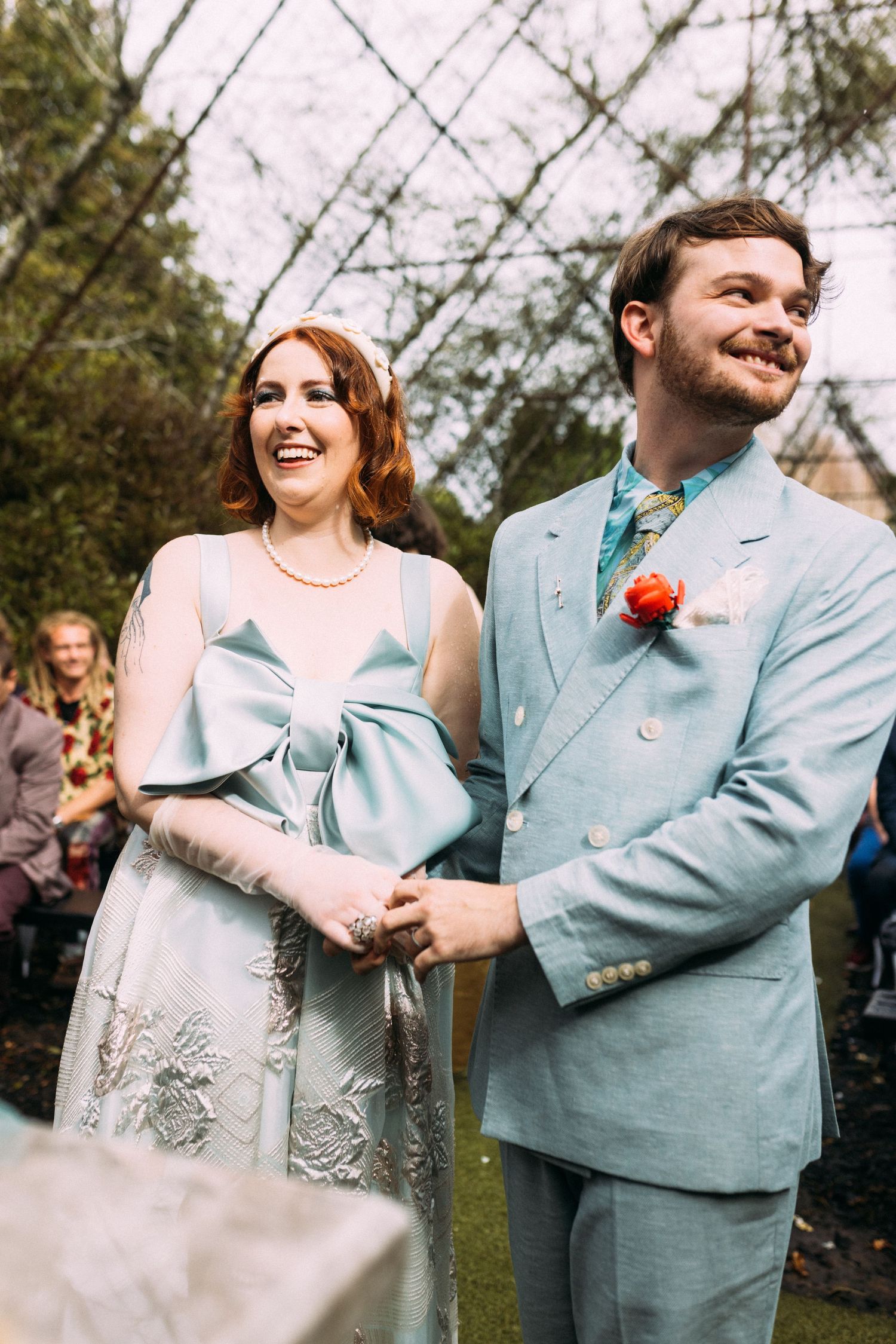 Laughing bride in light blue floral dress and groom in matching suit holding hands at outdoor ceremony.