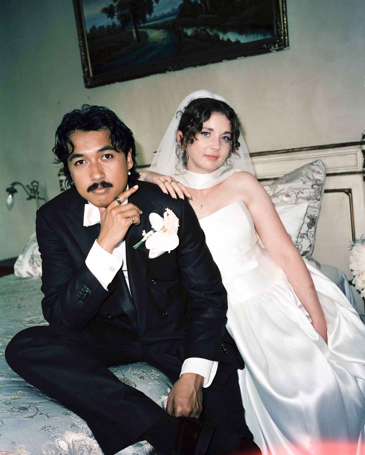 Bride and groom pose on ornate bed in vintage-style wedding portrait on film.