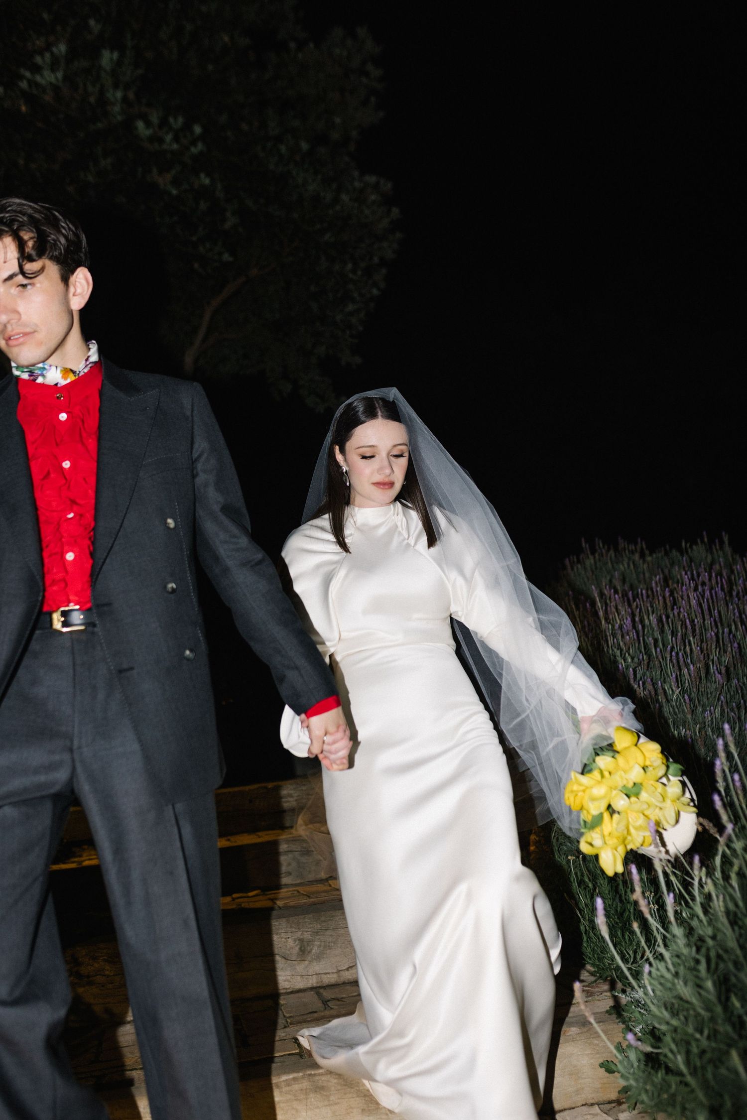 Bride in white satin gown with veil holds yellow bouquet while walking with groom at night outdoors.
