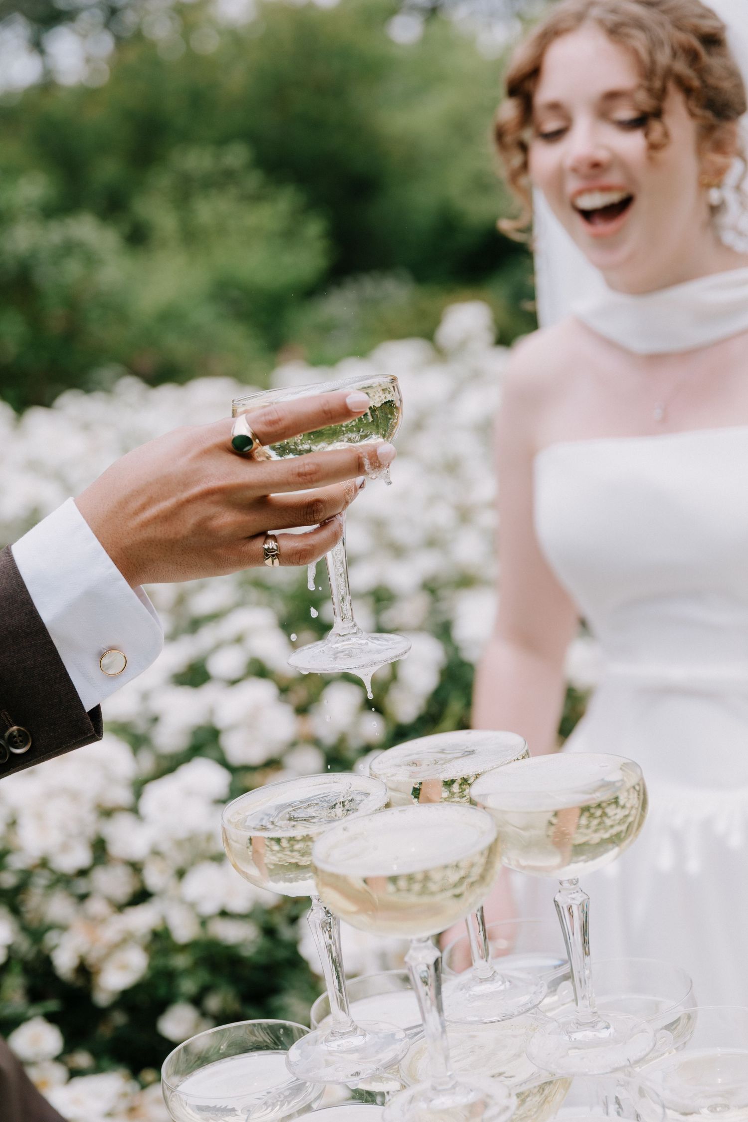 Bride laughs as champagne tower is served at outdoor wedding reception with white roses.