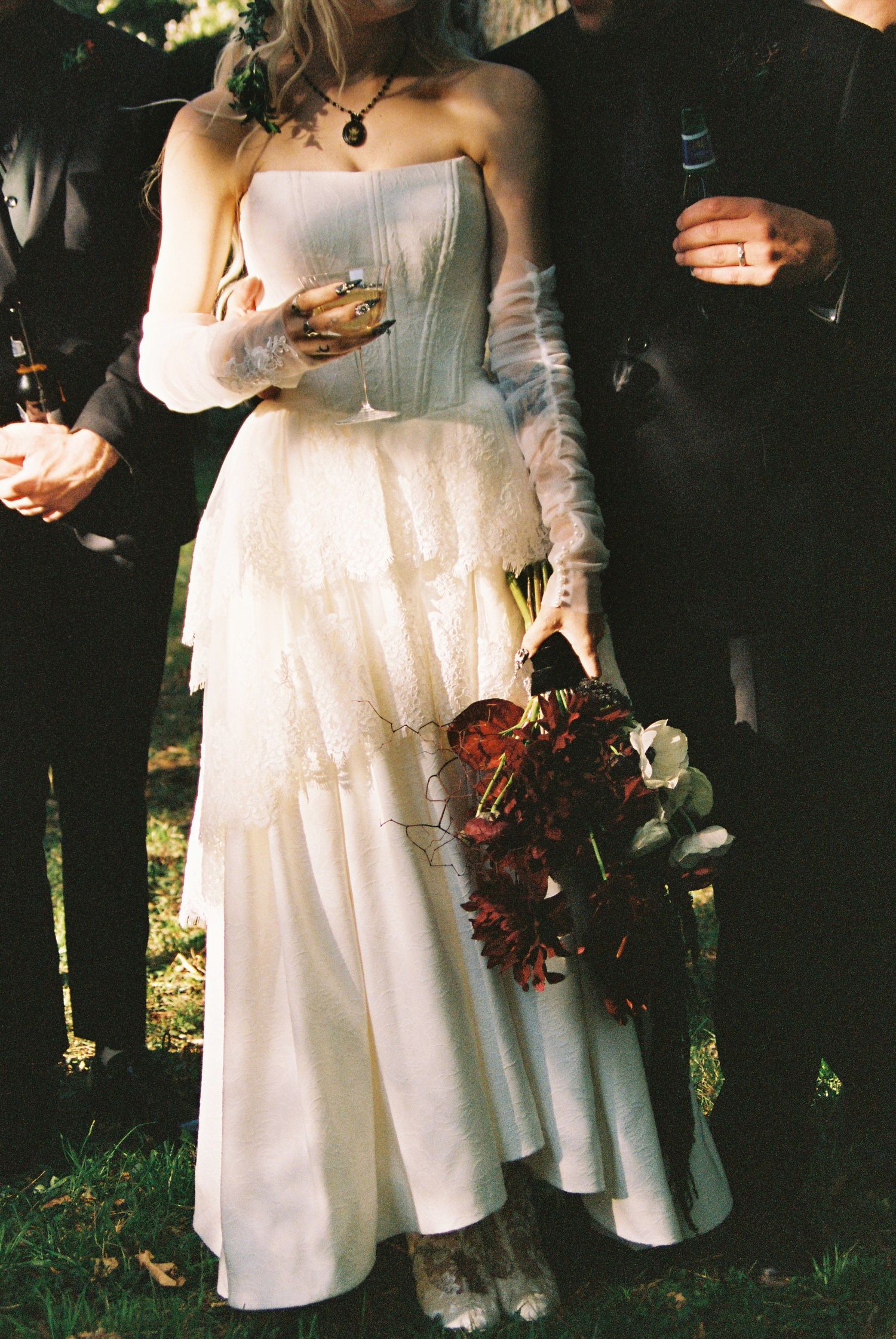 Bride in strapless white corset tiered gown holds dark red bouquet at an outdoor wedding celebration.