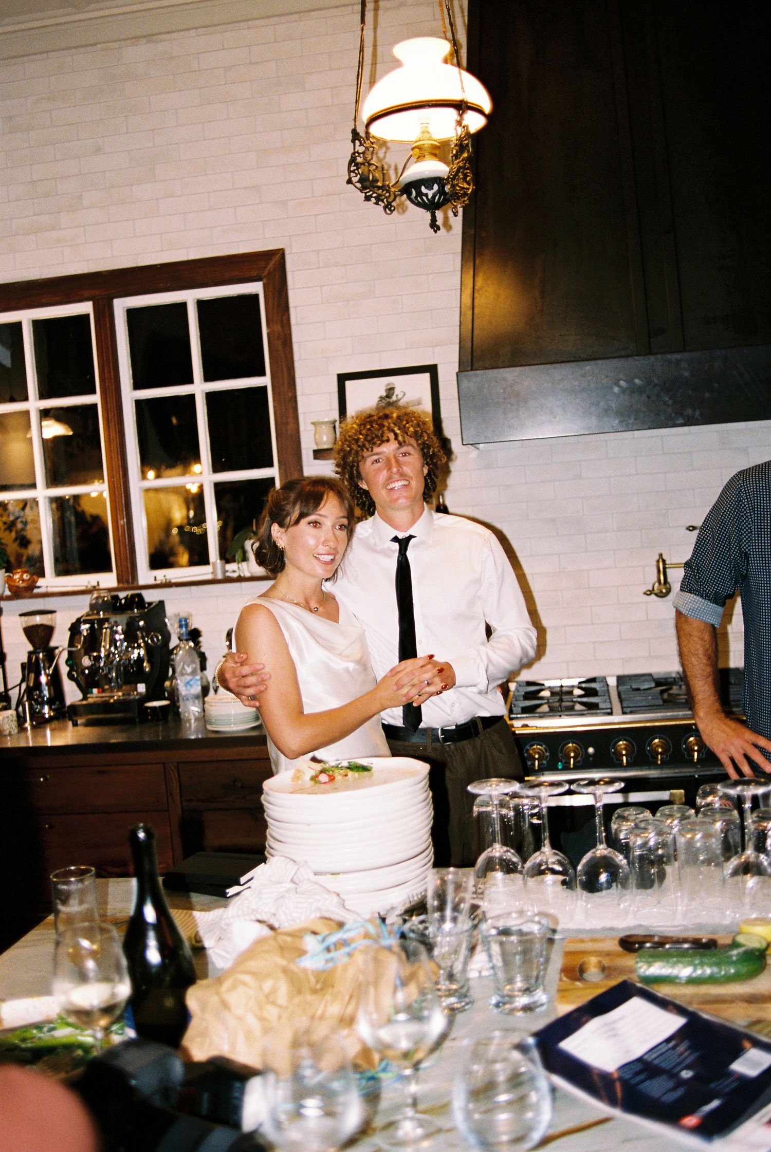 Bride and groom with curly hair cutting their wedding cake together in a rustic kitchen setting.