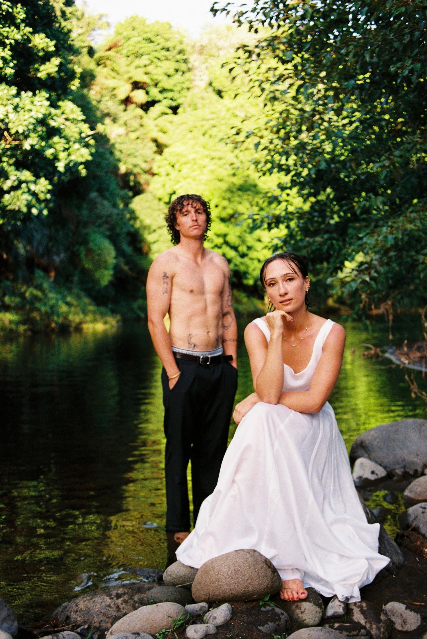 Couple posing by a lush green river, woman in white dress seated on rocks, man standing.