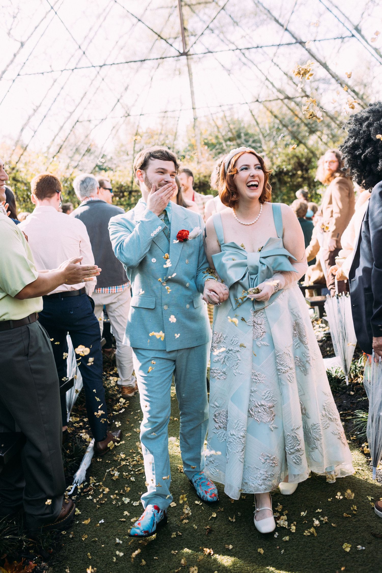 Joyful newlyweds walk down aisle in matching light blue outfits as confetti falls around them.