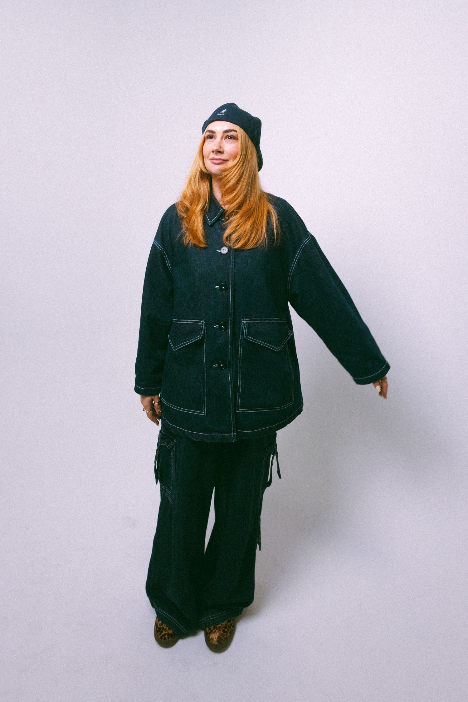Woman in dark denim jacket, wide-leg pants, and beanie hat posing against white background.