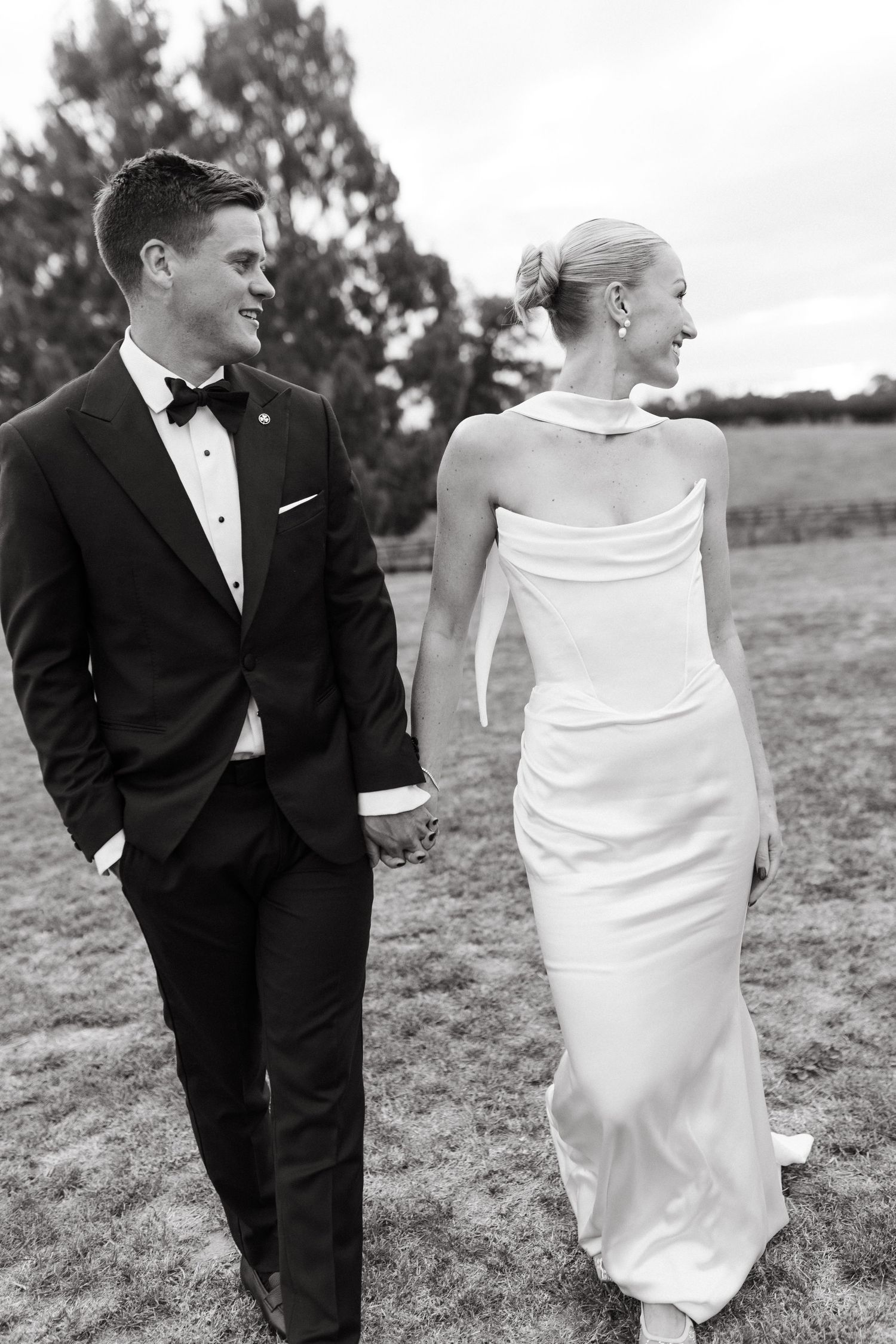 Black and white photo of bride in backless gown and groom in tuxedo holding hands walking in field.