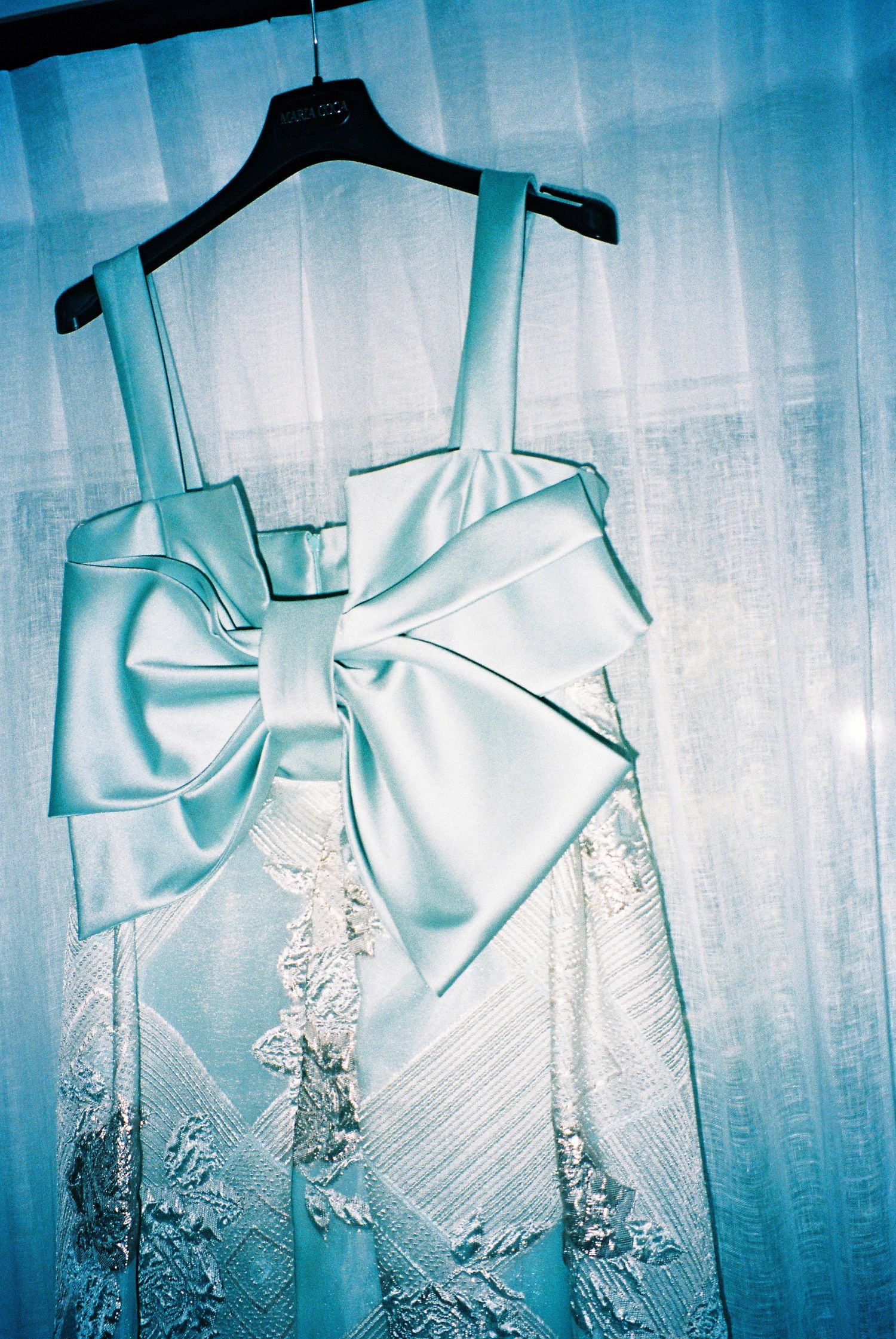 Light blue wedding dress with large satin bow detail hanging on black hanger against sheer curtain.