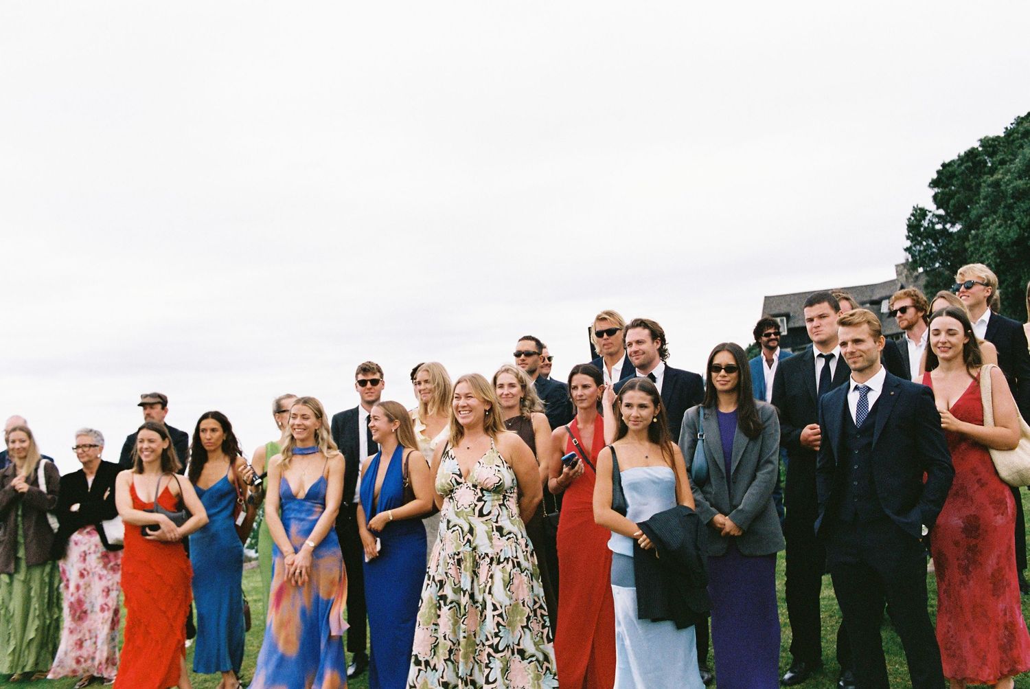 Large group of elegantly dressed wedding guests pose together on an outdoor lawn.