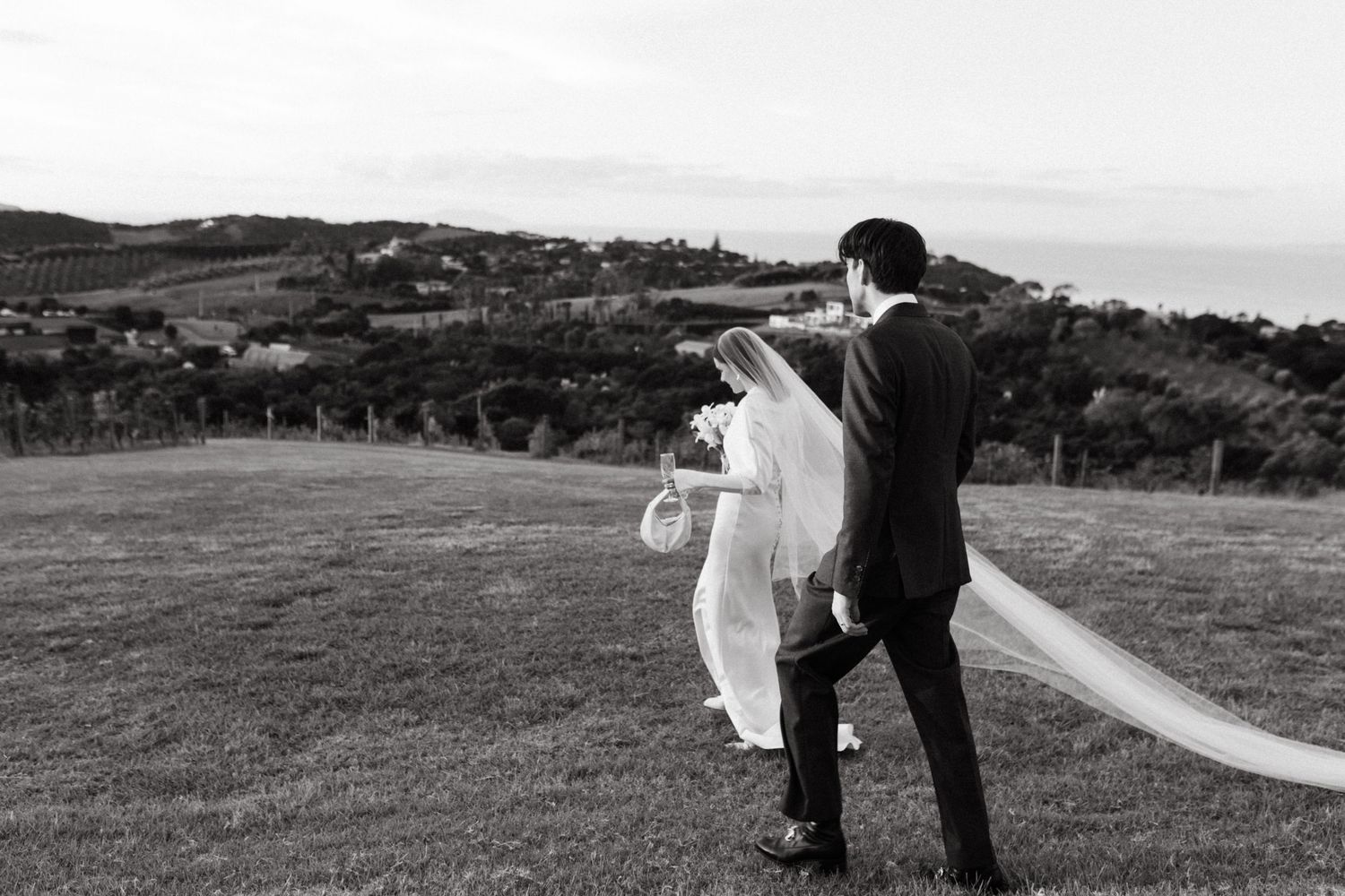 Black and white photo of bride and groom walking across grassy hillside, wedding veil flowing in the breeze.