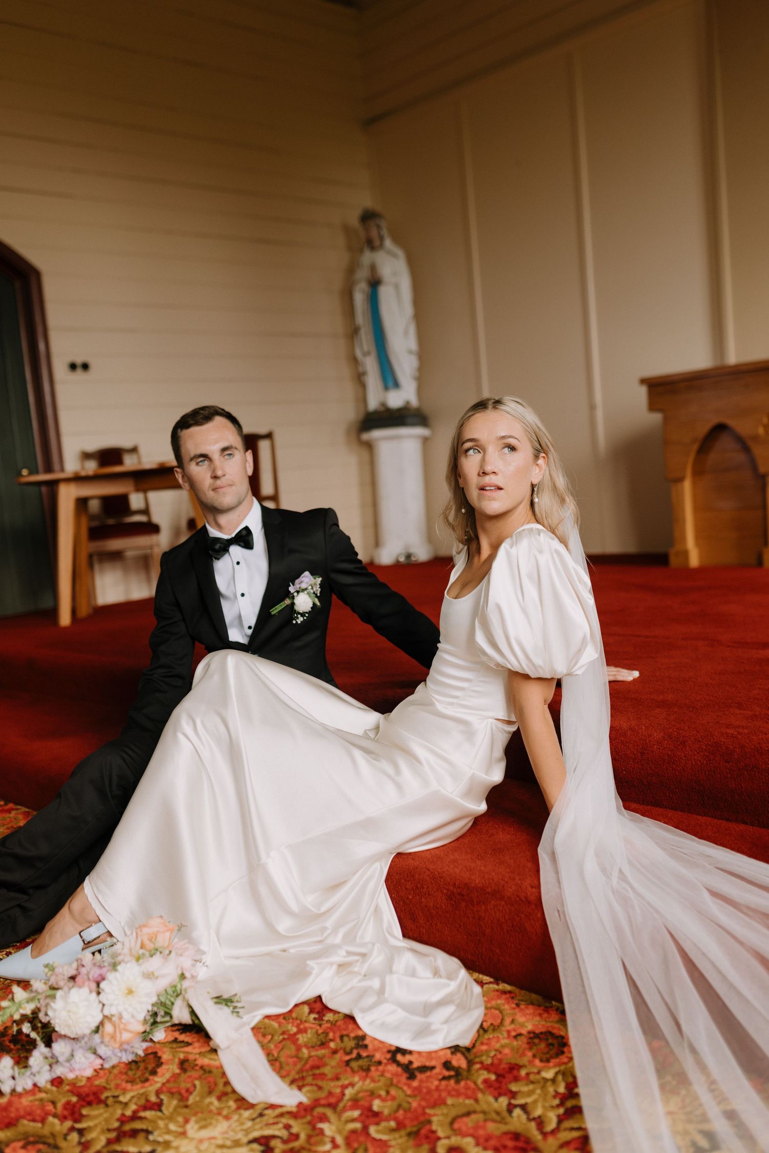 Bride and groom in formal attire seated on red carpet inside a classic wooden chapel interior.