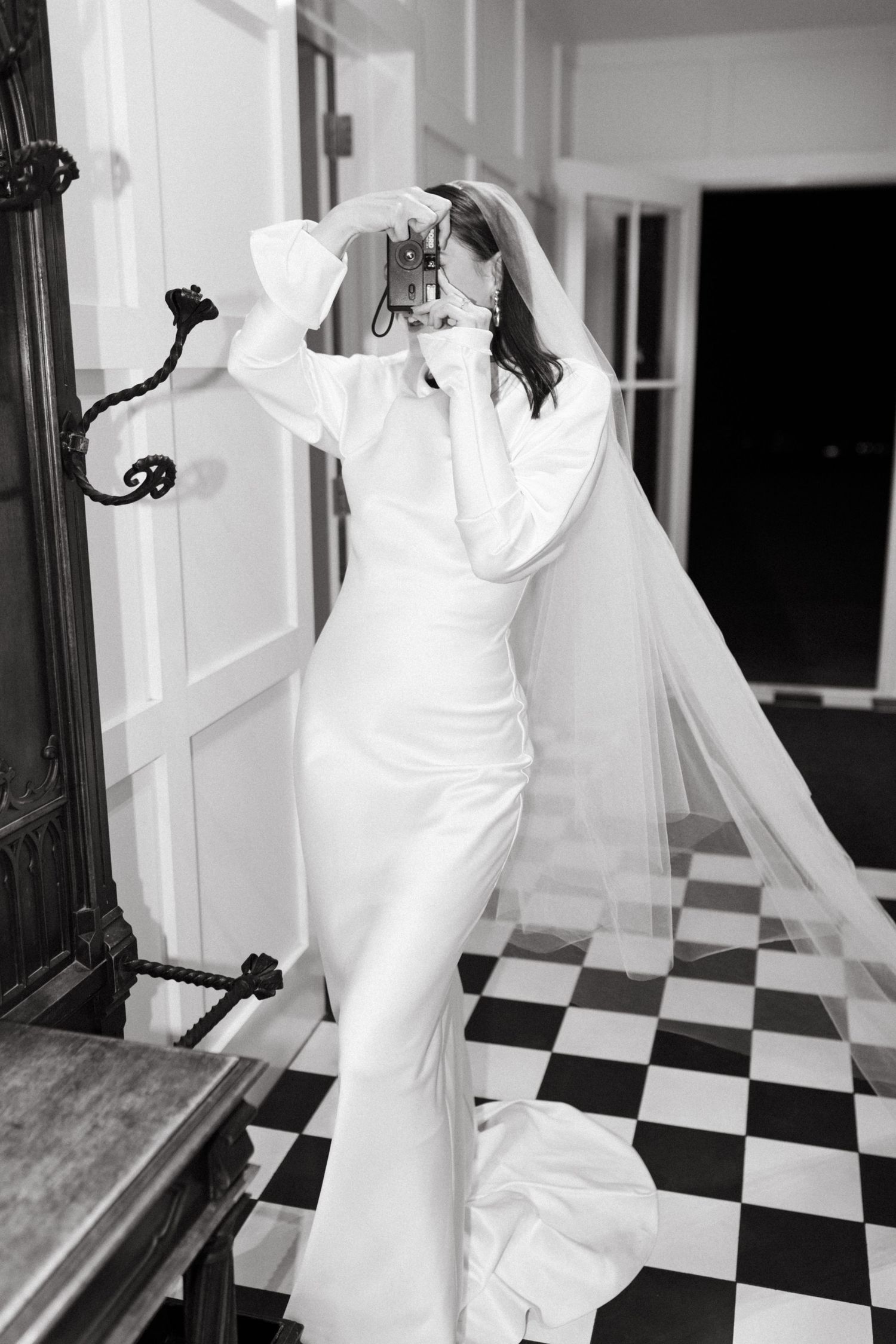 Black and white photo of bride in white satin gown and veil on checkered floor adjusting her veil.
