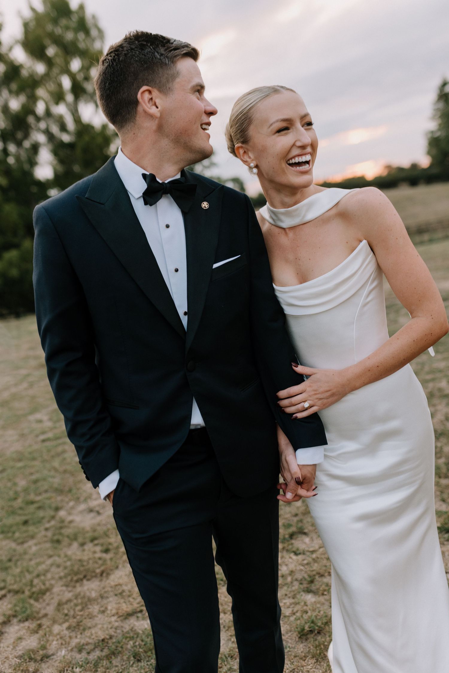 Smiling bride and groom in black tuxedo and white cowl-neck wedding dress at sunset outdoor ceremony.