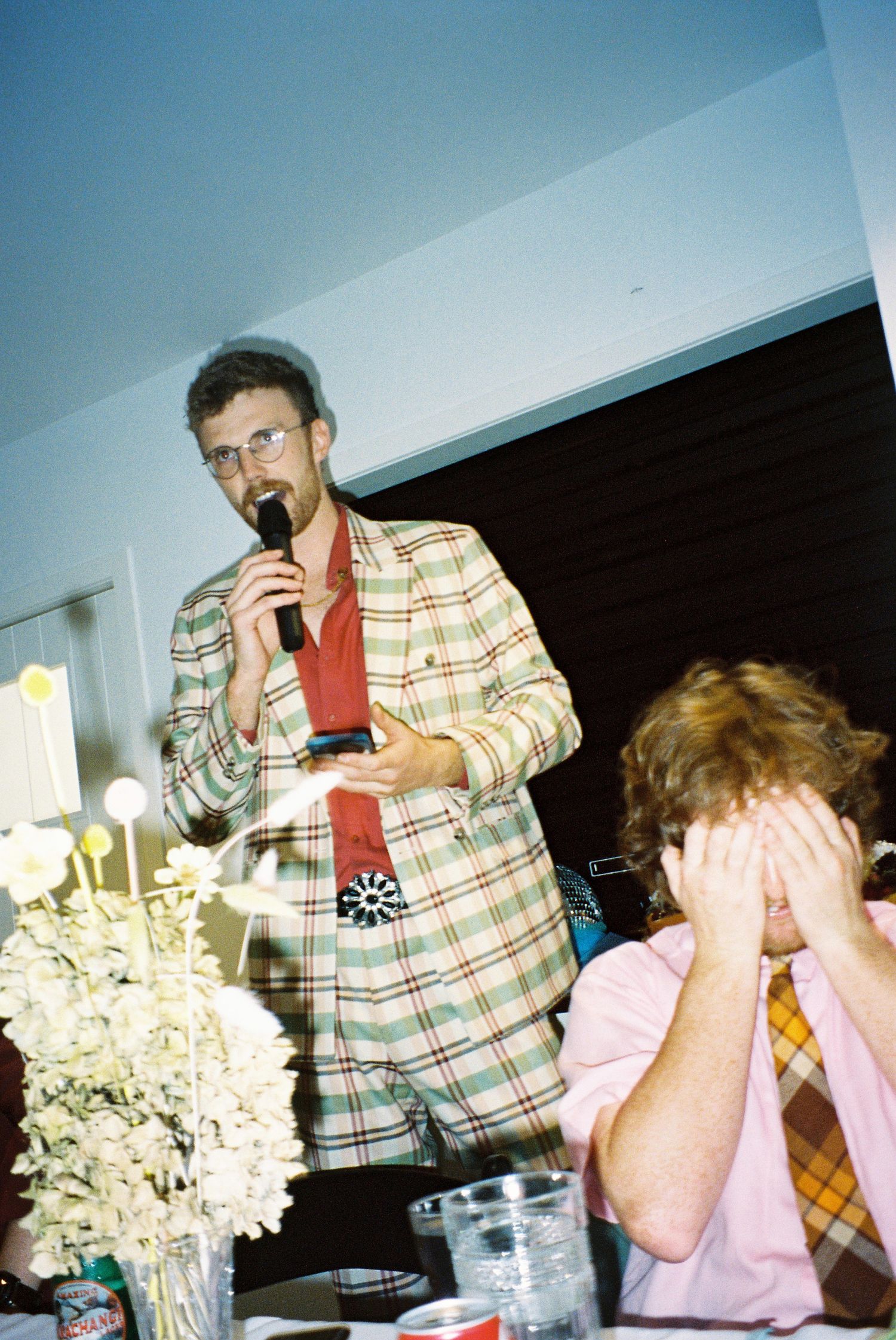 Man in plaid suit speaks into microphone at wedding reception while guest covers face nearby.