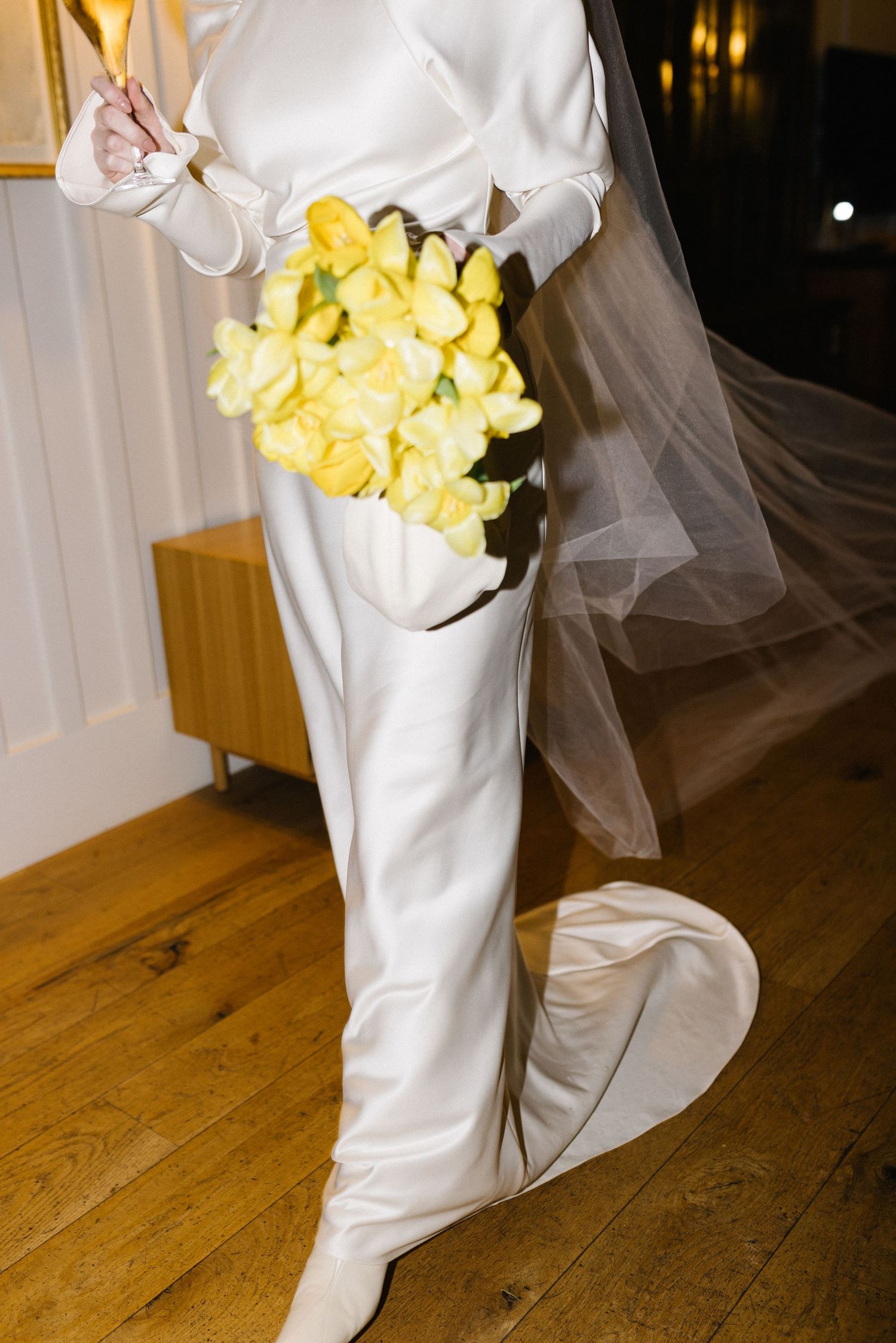 Close-up of bride in white satin gown holding yellow tulip bouquet with veil flowing behind her.