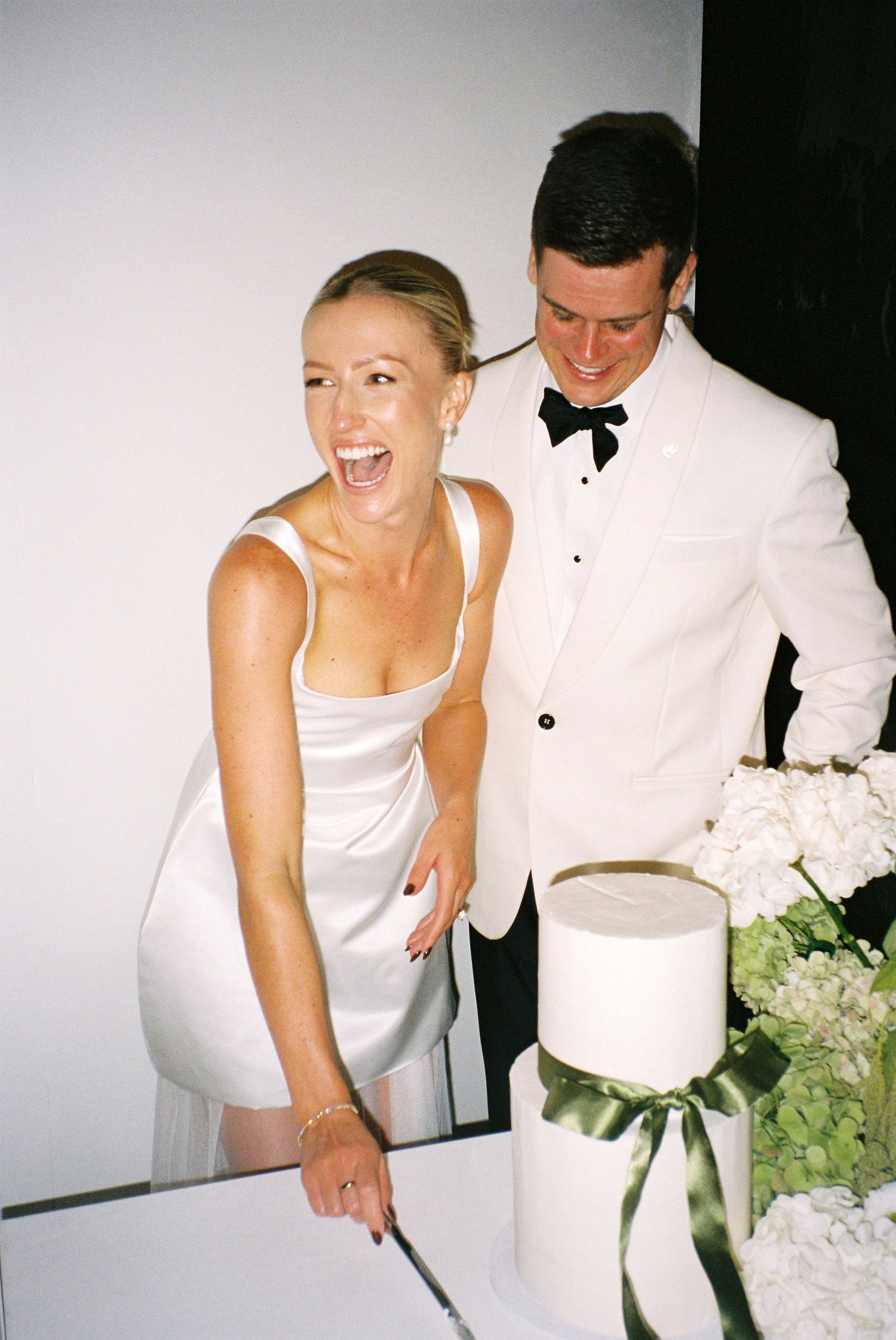 Laughing bride in white dress with groom in white tuxedo cutting tiered wedding cake with green ribbon.