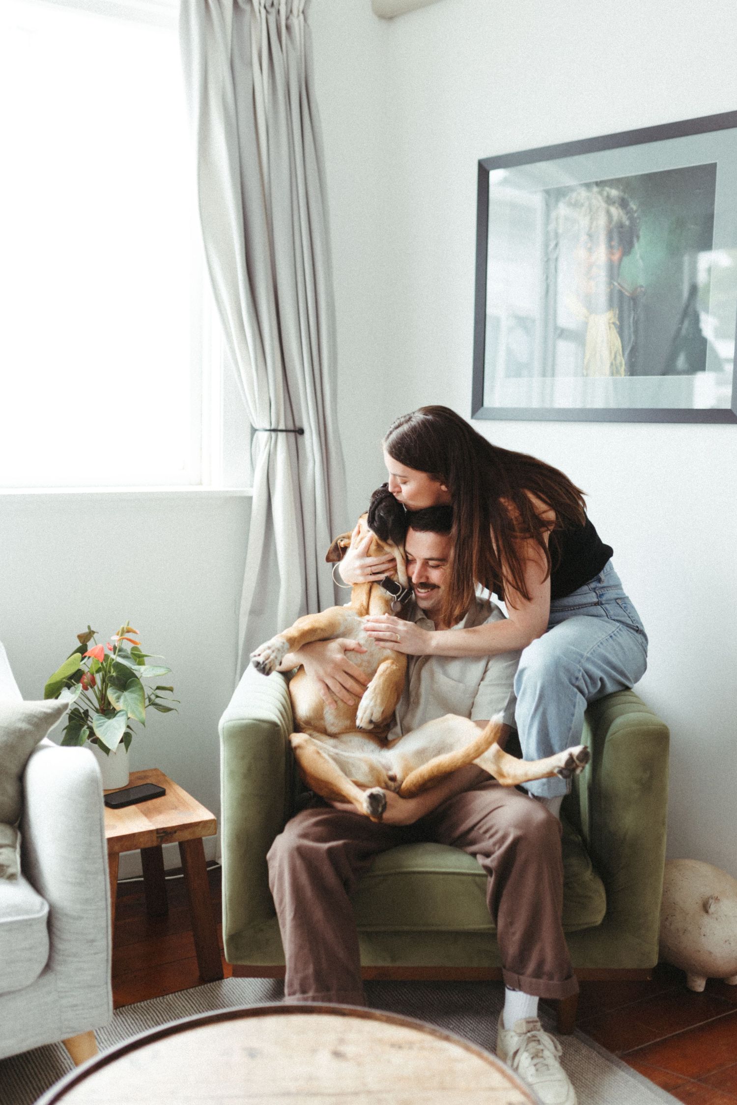 Happy couple cuddles with their dog on a green armchair in a bright cozy living room.