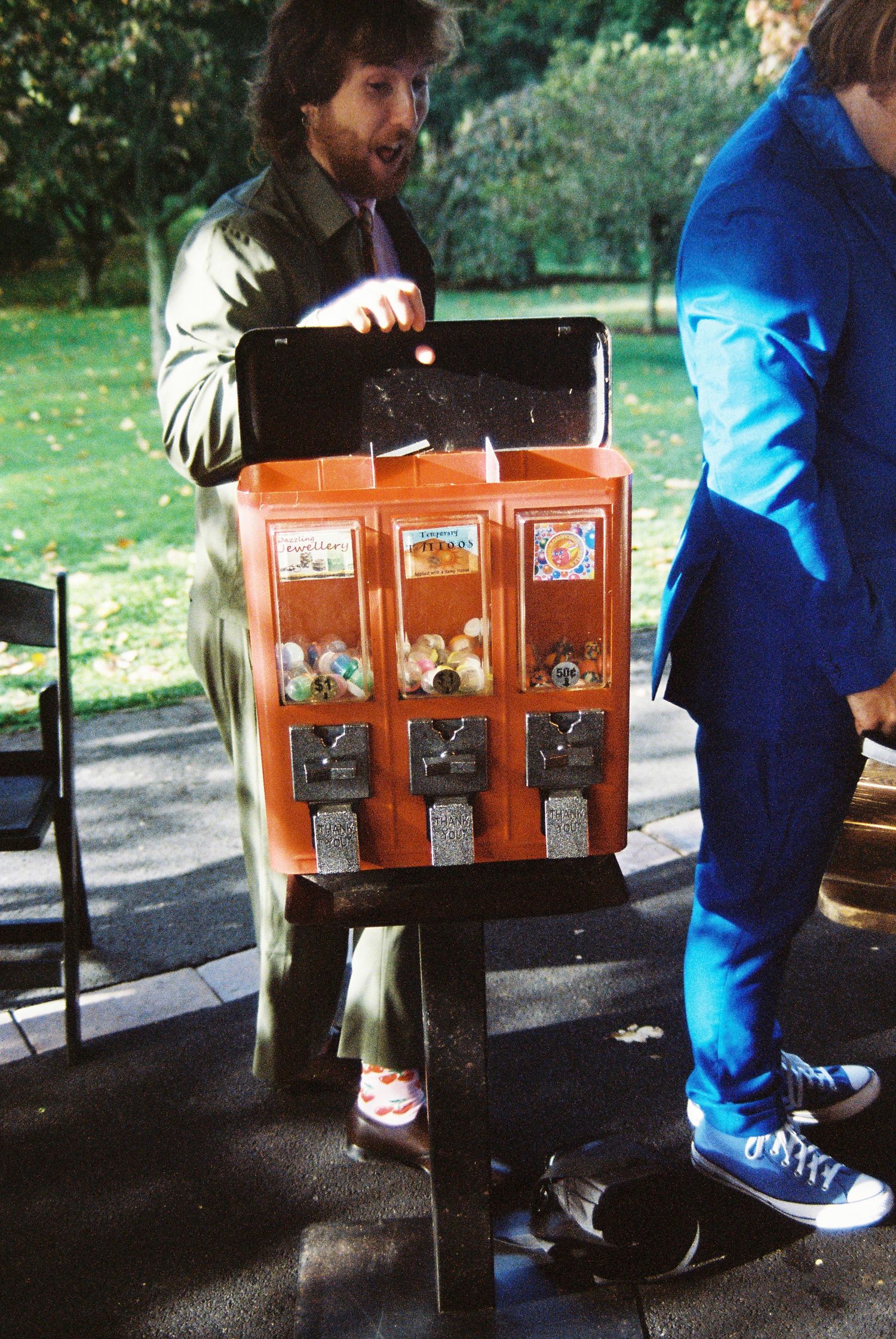Man operates vintage gumball machine at outdoor wedding reception beside guest in blue suit.