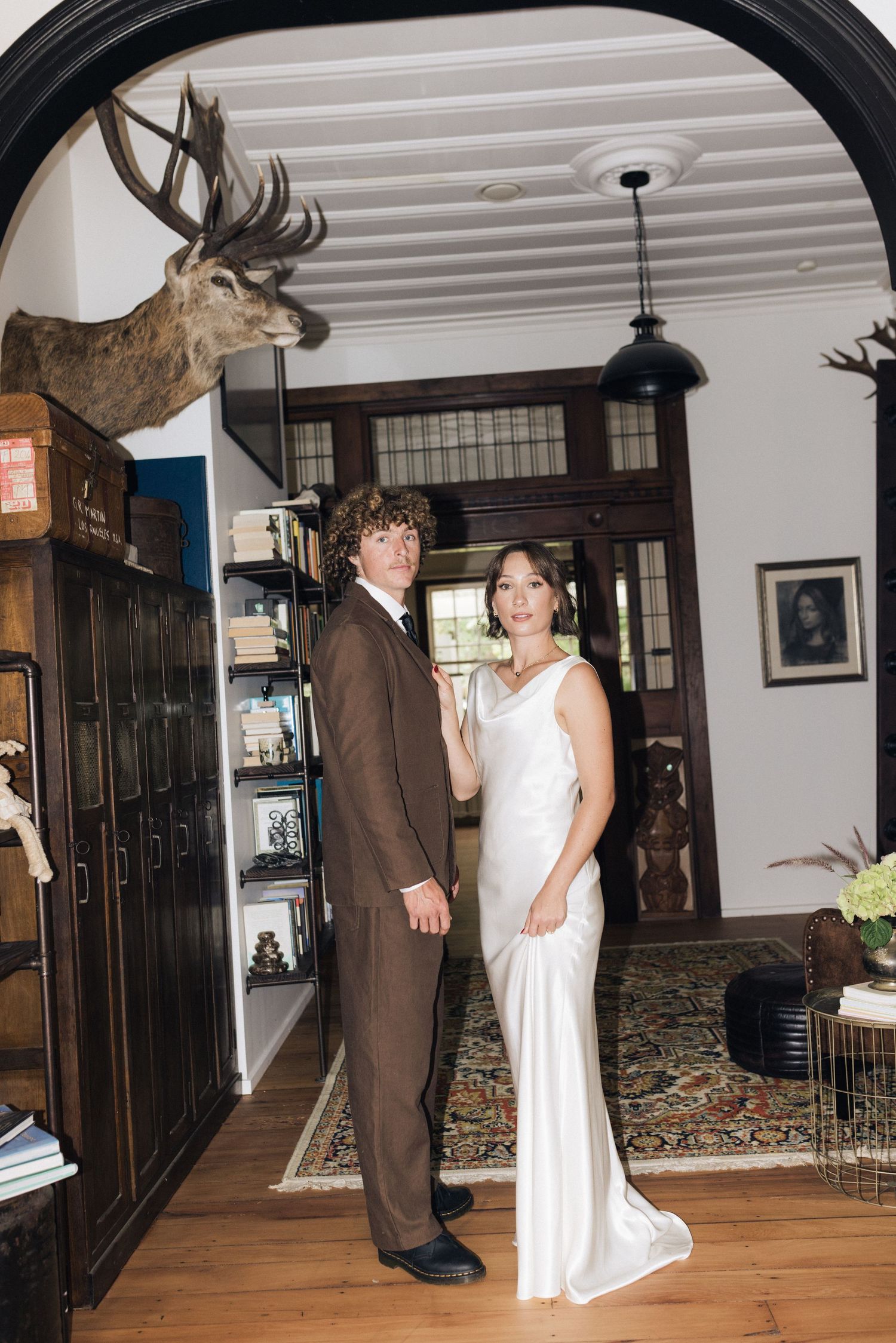 Bride in white gown and groom in brown suit posing under archway with mounted deer head.