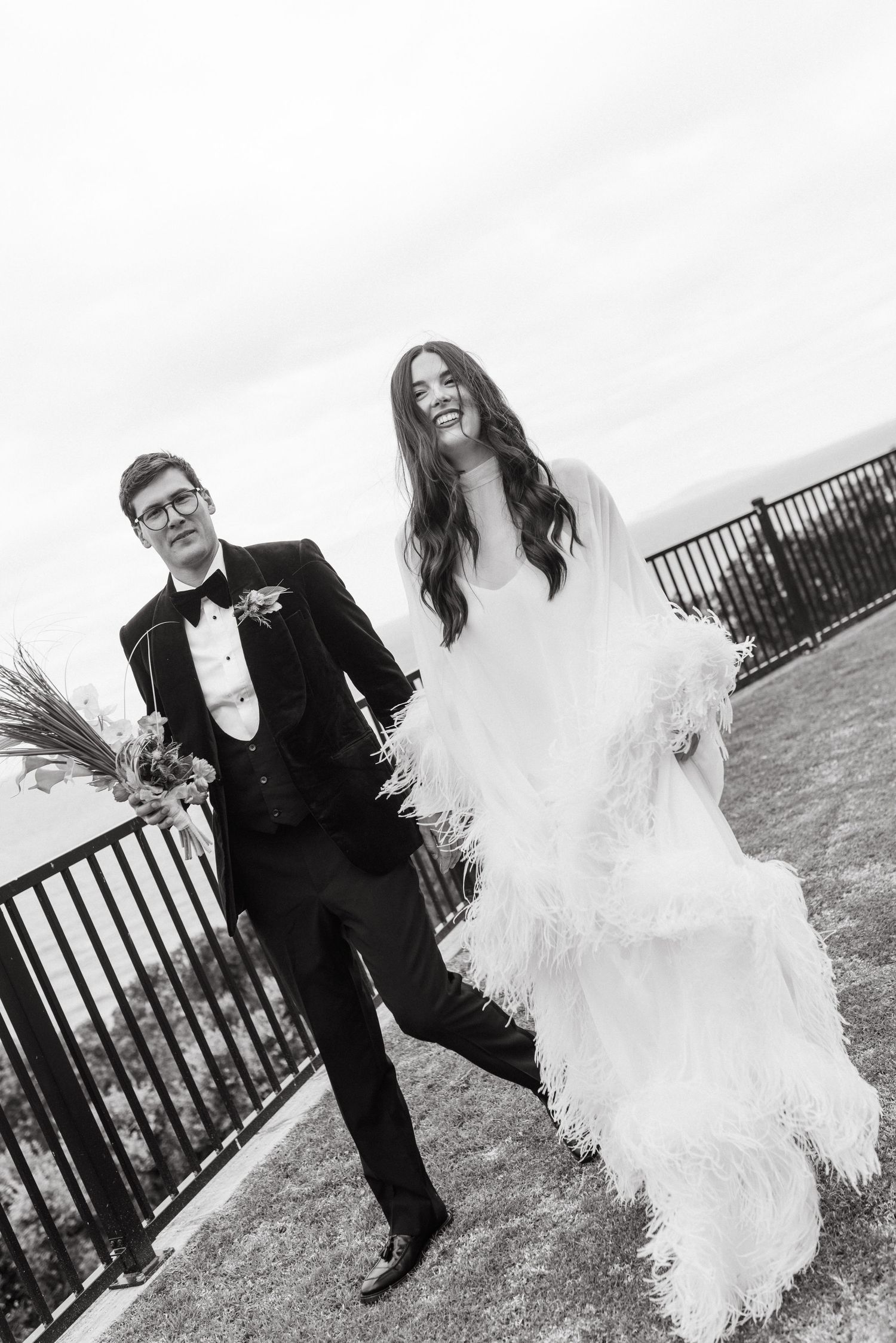 Black and white photo of laughing bride in feathered gown and groom in tuxedo walking outdoors at wedding.