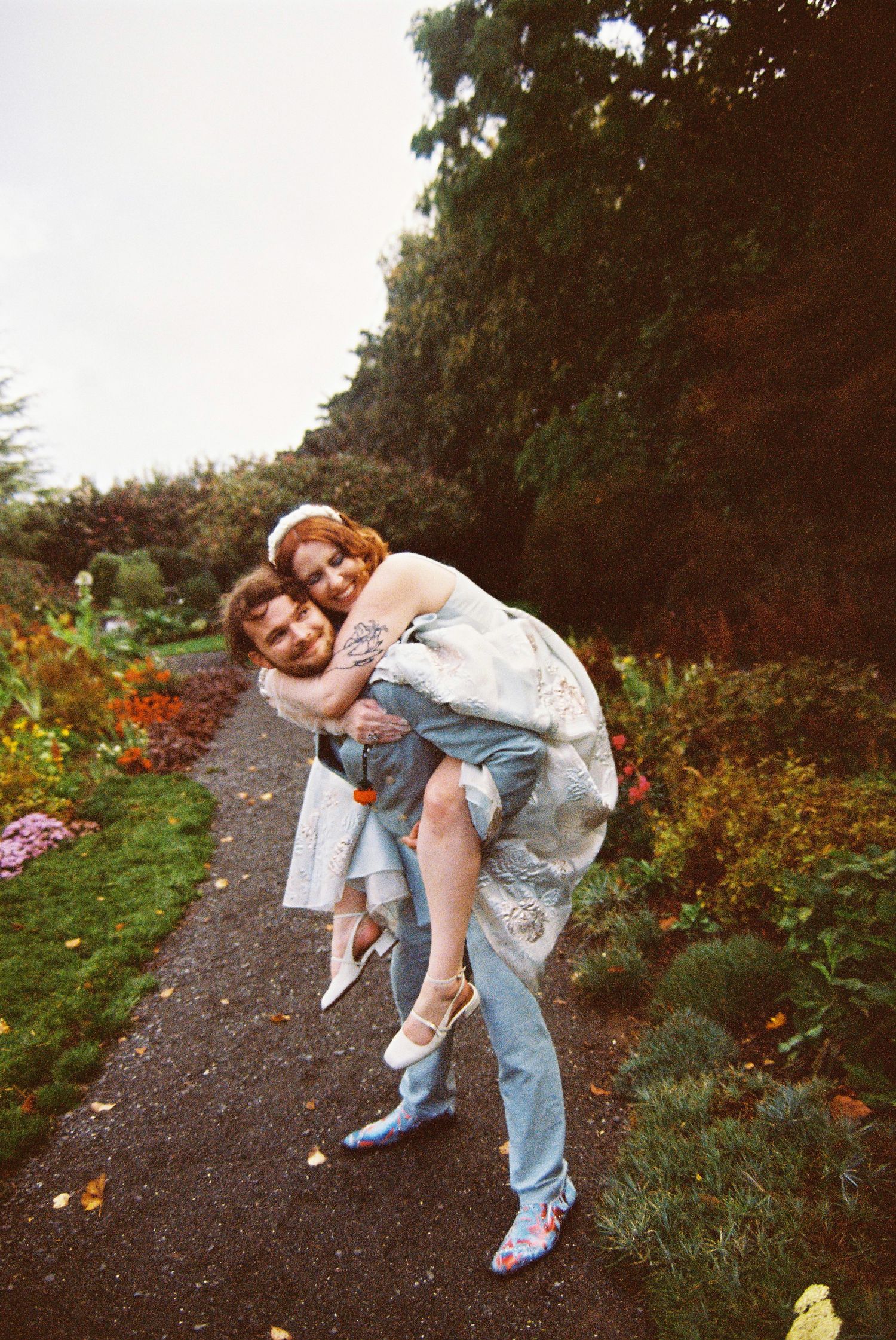 Groom gives bride a piggyback ride along a garden path lined with colorful flowers.