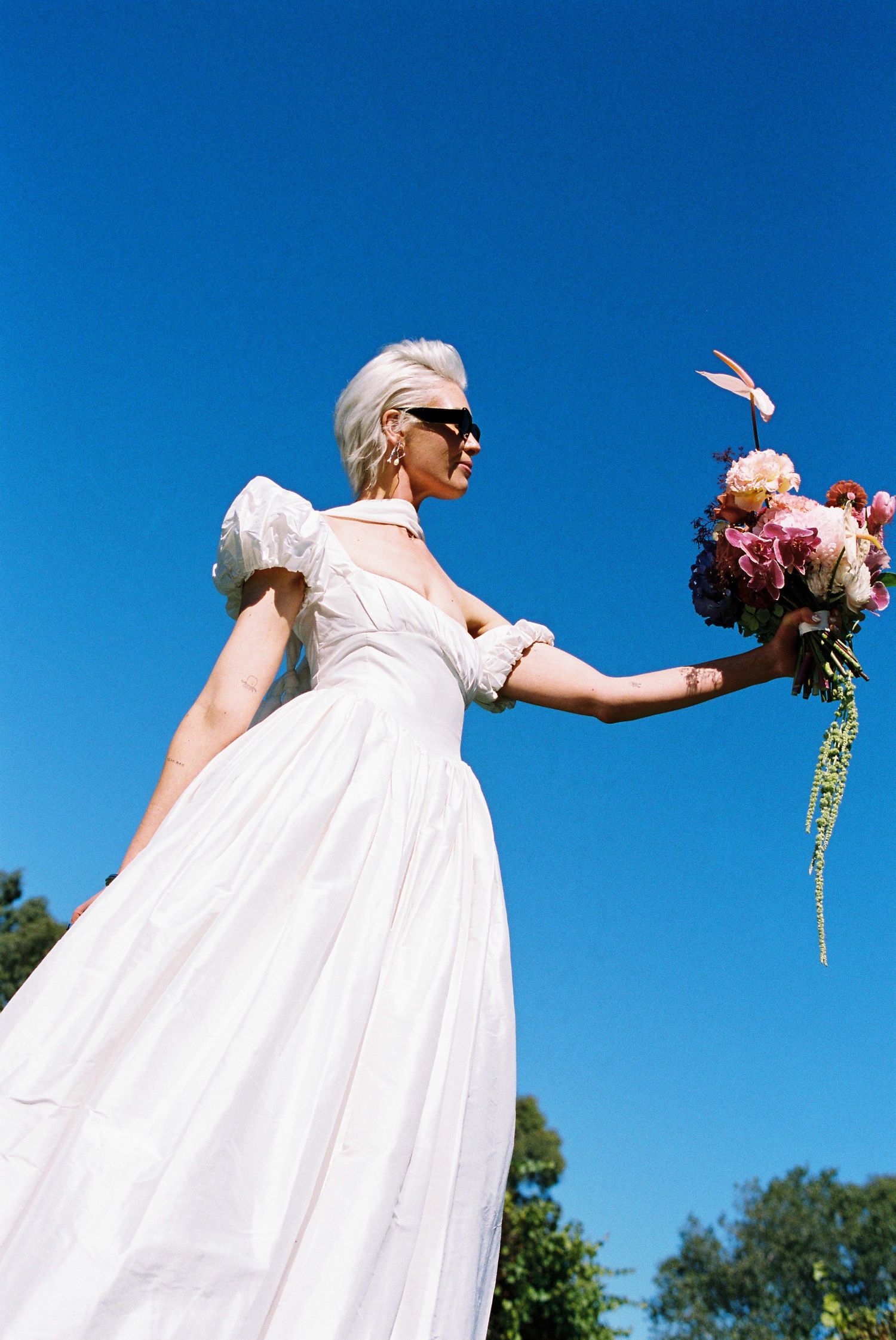 Bride in white puff-sleeve gown holding pink bouquet against a vivid blue sky.