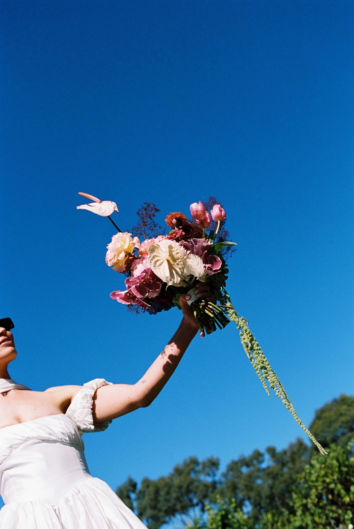 Bride in white dress raising colorful floral bouquet with pink roses against bright blue sky.