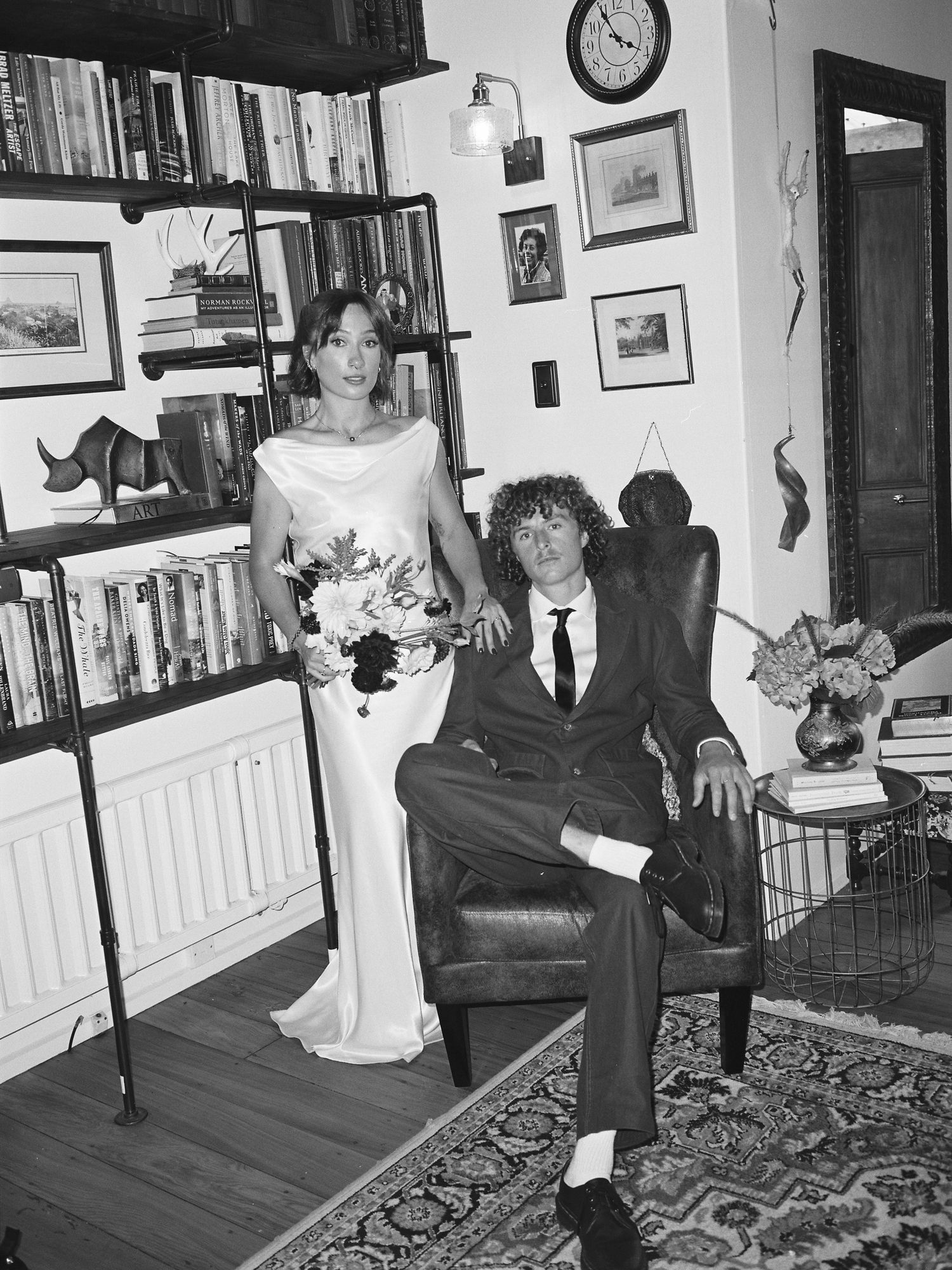 Black and white portrait of bride and groom posing in cozy bookshelf-lined room with floral bouquet.