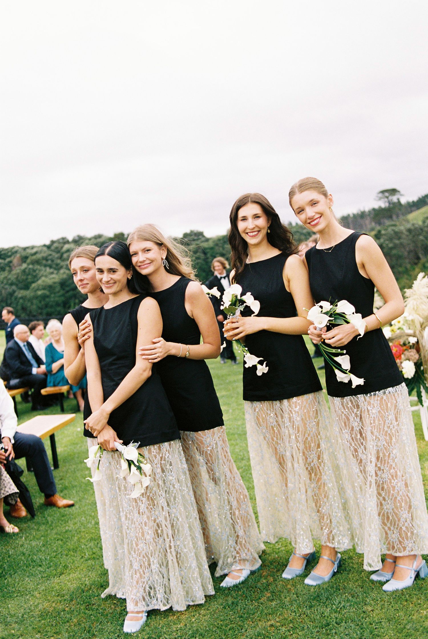 Four bridesmaids in matching black tops and lace skirts holding white bouquets at outdoor wedding ceremony.