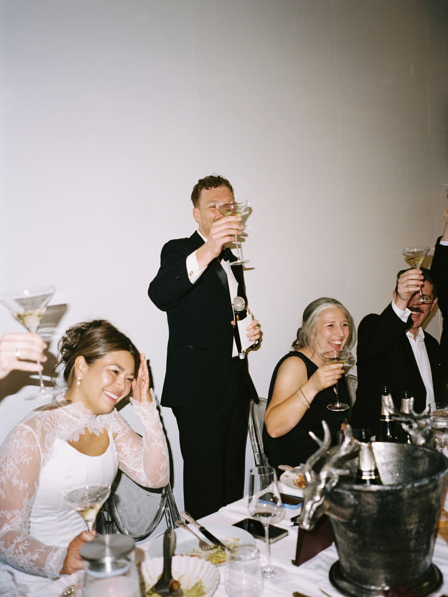 Wedding guests raising champagne glasses in a toast at indoor reception, bride smiling in lace gown.
