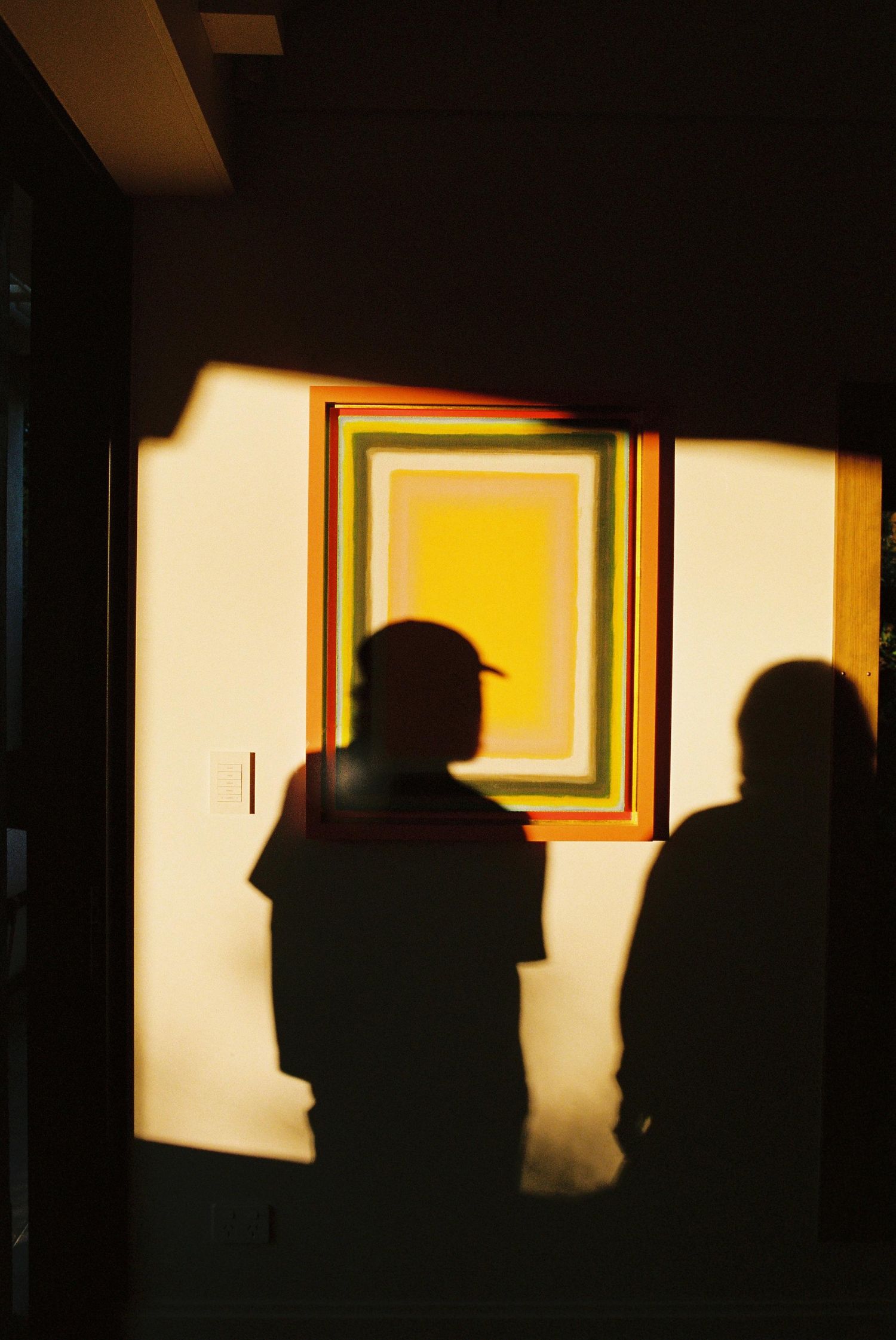 Silhouettes of two people cast against a wall beside a vibrant yellow geometric framed artwork.