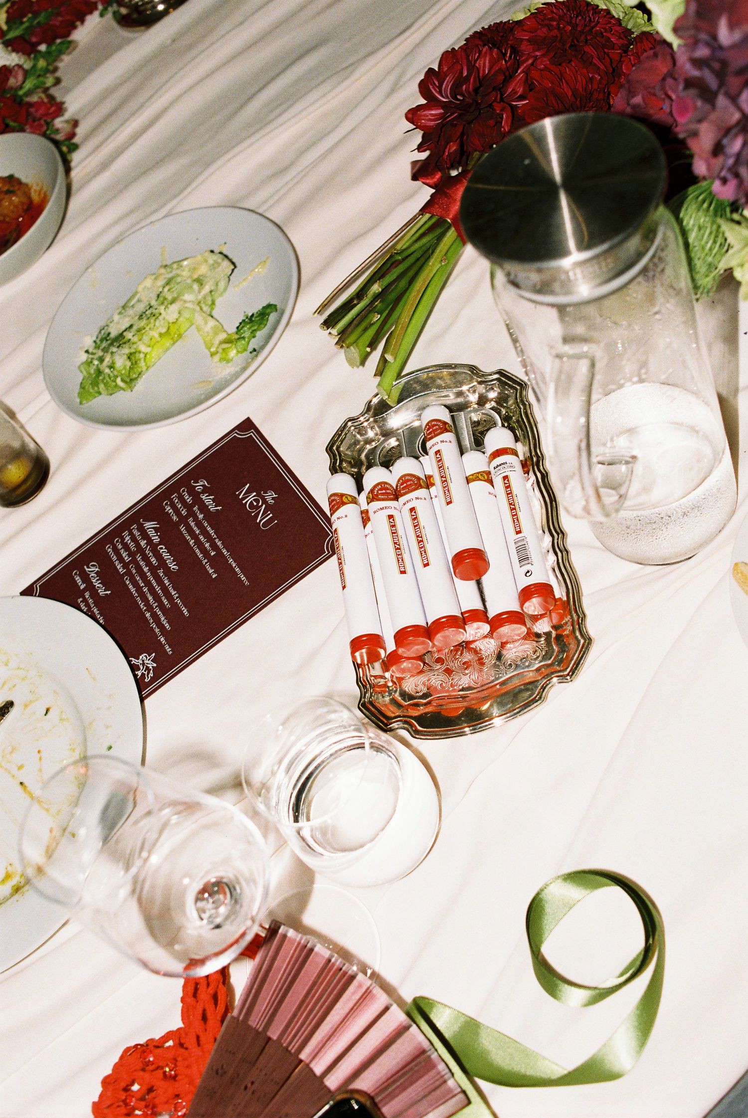 Elegant wedding table setting with dark red flowers, menu card, shot tubes, and green ribbon.