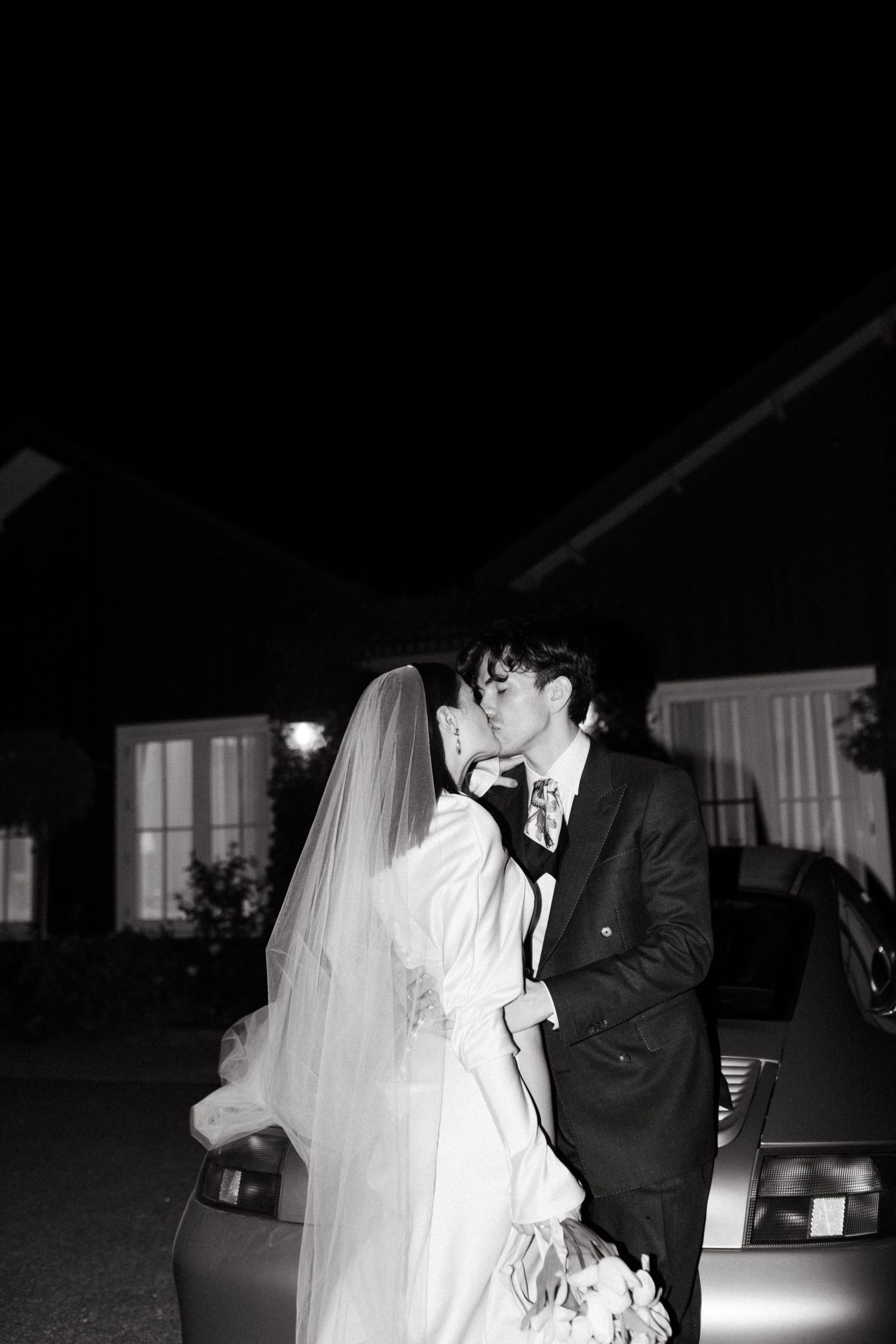 Black and white photo of bride and groom sharing a kiss beside a car at night after their wedding.