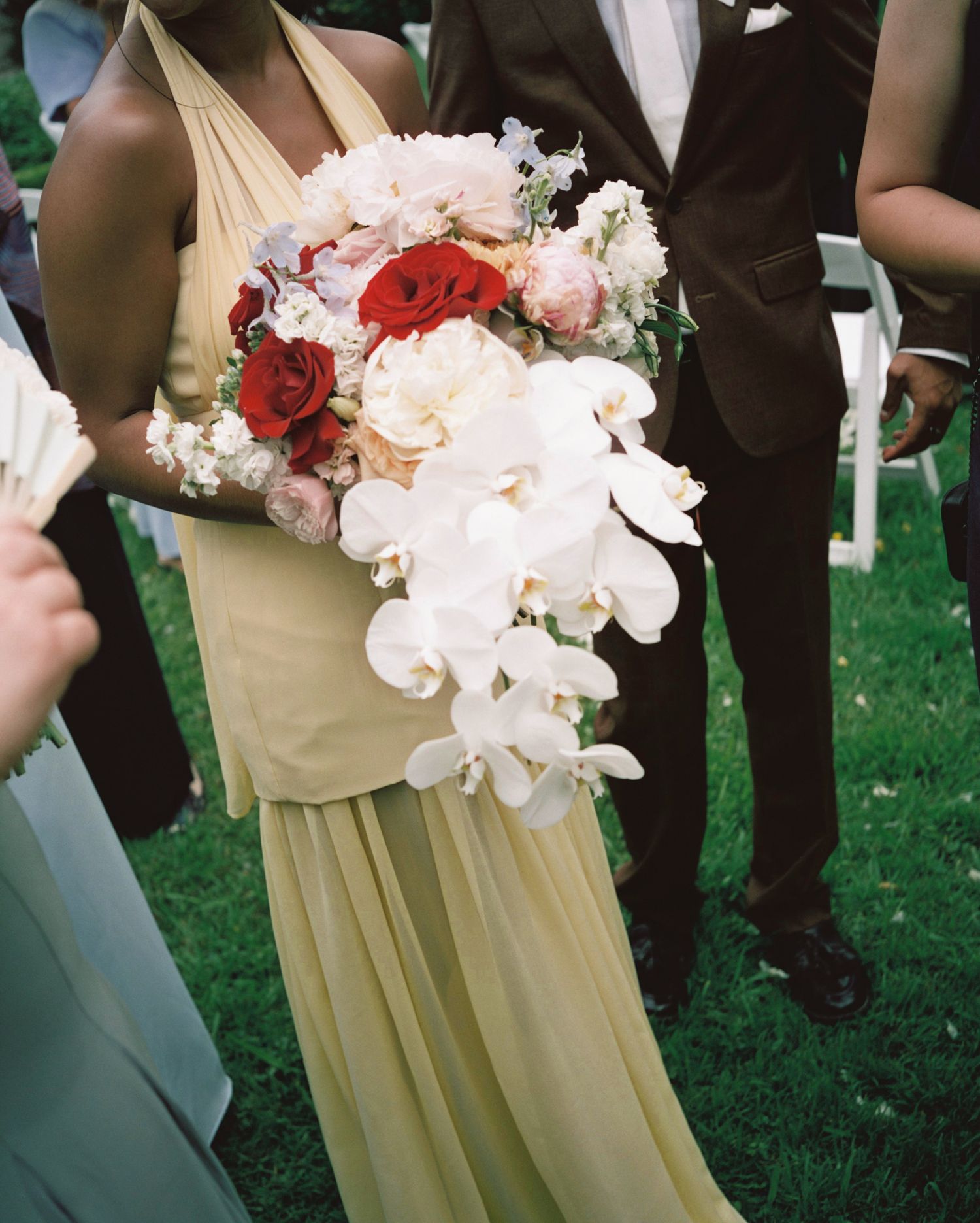 Bridesmaid in yellow gown holds cascading bridal bouquet with white orchids and red roses at outdoor wedding ceremony.