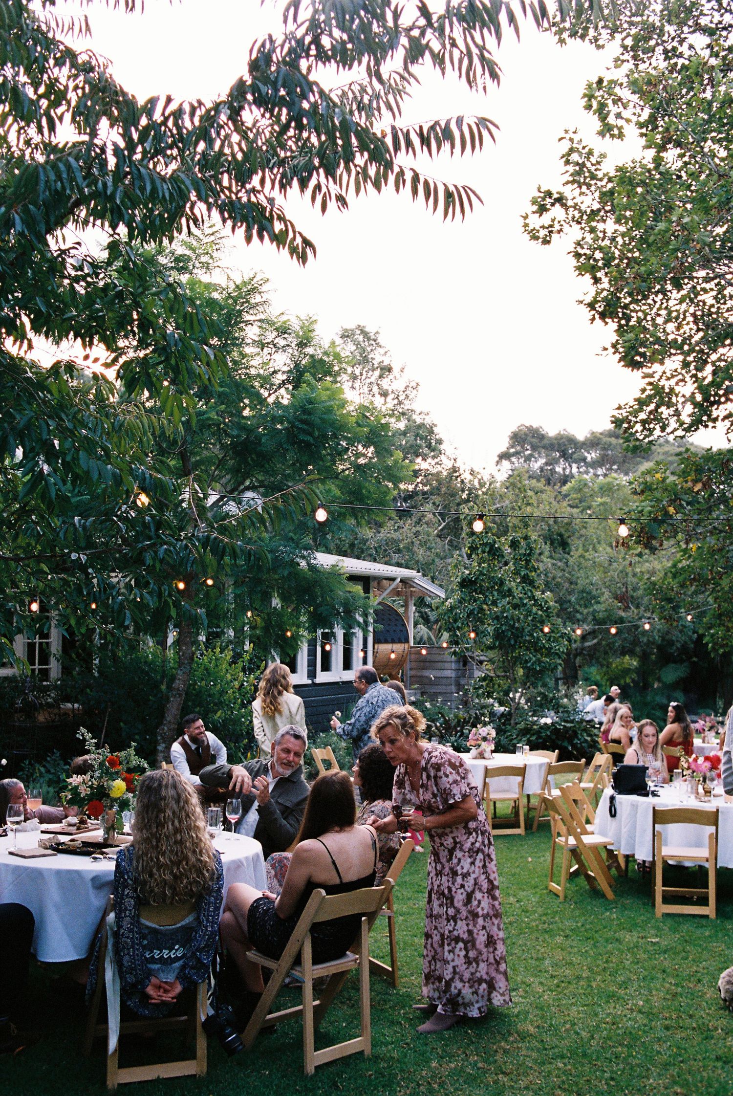 Outdoor garden wedding reception with guests dining at white-clothed tables under string lights and trees.